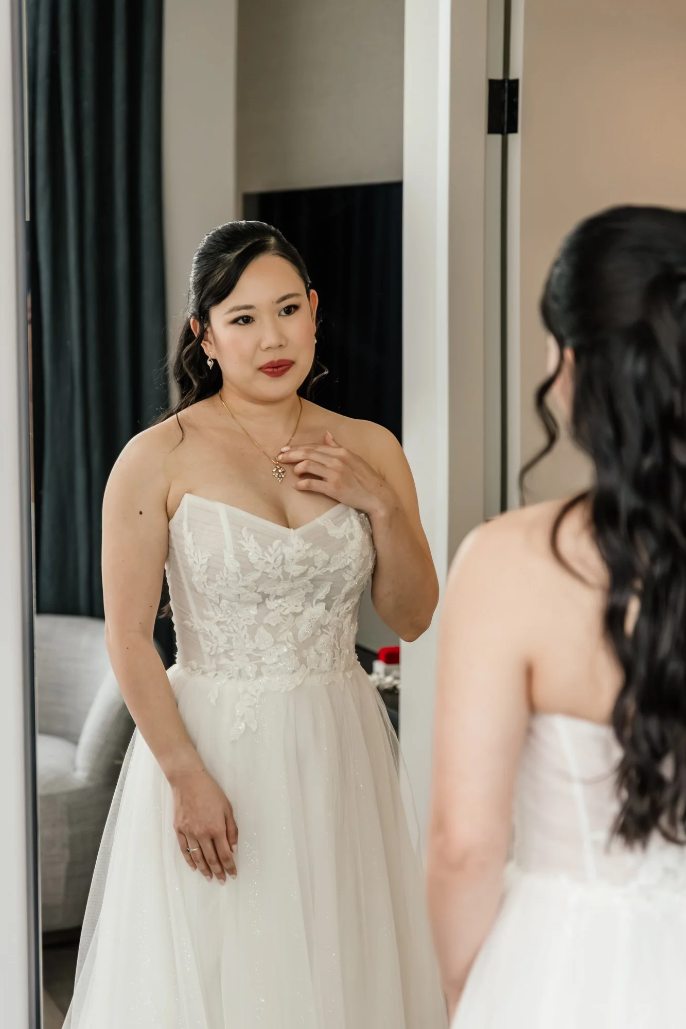 A woman in a wedding dress looking at herself in the mirror, adjusting her dress with her hand near her neck, with hair and makeup done for her wedding.