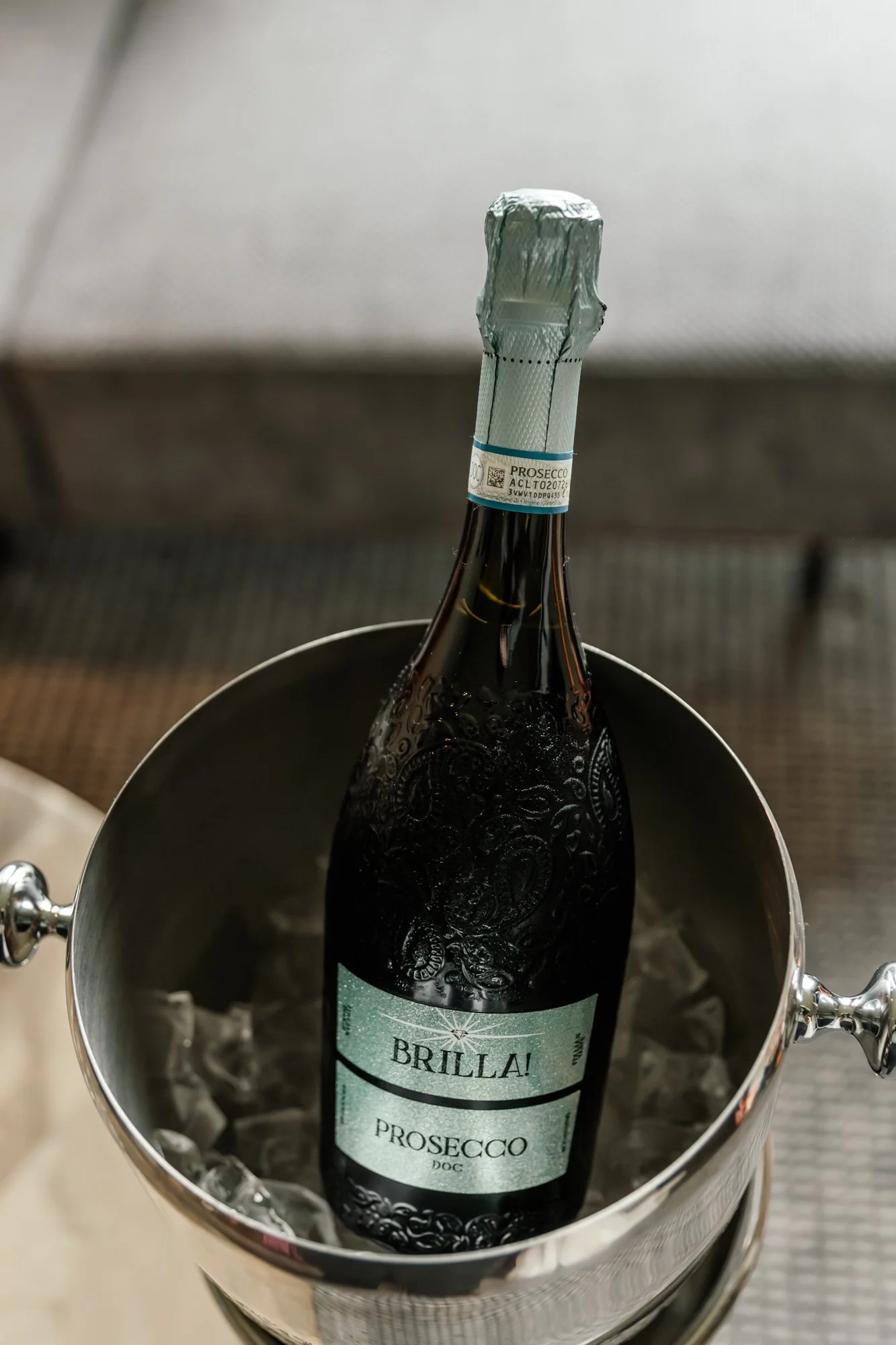 A bottle of sparkling wine or champagne with a blue and white label reading 'BRILLA! PROSECCO' is displayed inside a silver ice bucket filled with ice, on a countertop.