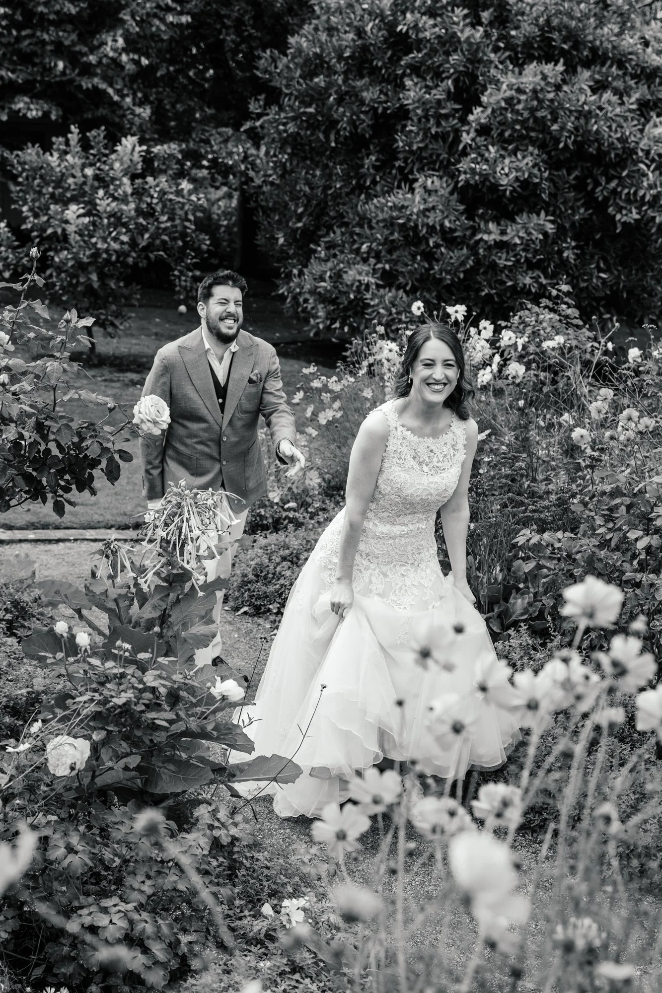 Black and white photo of a bride and groom in a garden, with the bride smiling and holding her wedding dress, and the groom smiling and walking behind her among flowers and bushes.