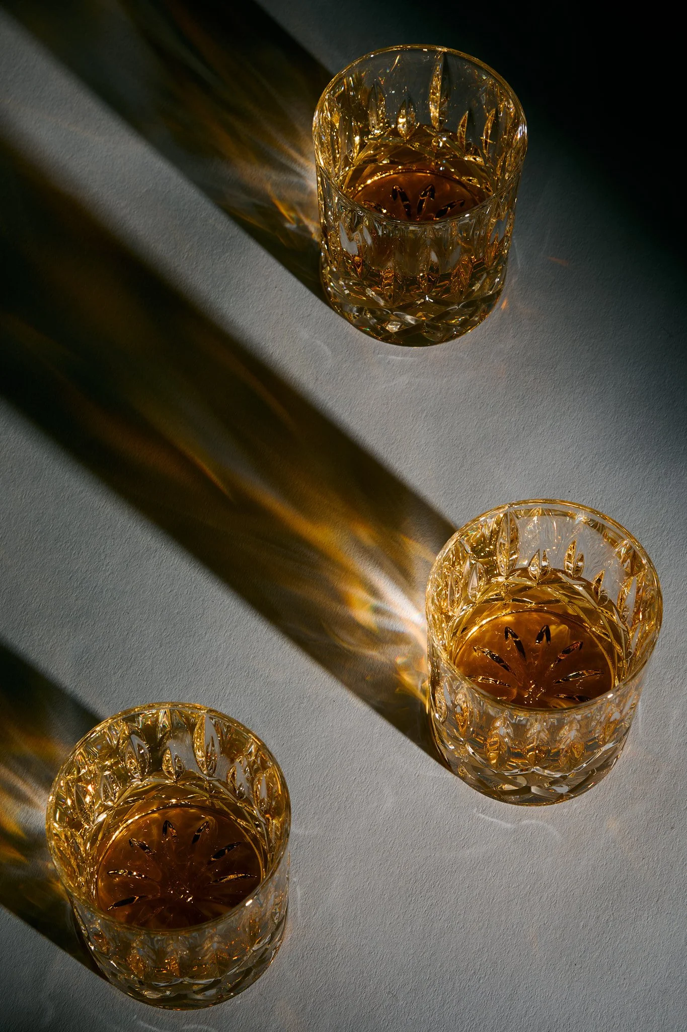 Three crystal glasses filled with a golden-colored liquid, placed on a light-colored surface, with light casting reflections and shadows.