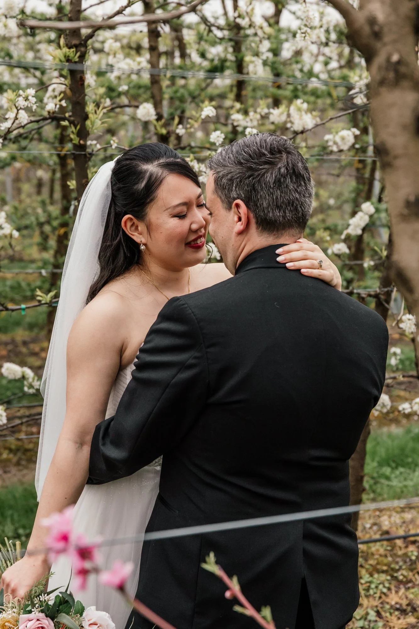 A bride and groom sharing a close moment, standing in a garden with blooming white flowers and green foliage in the background.