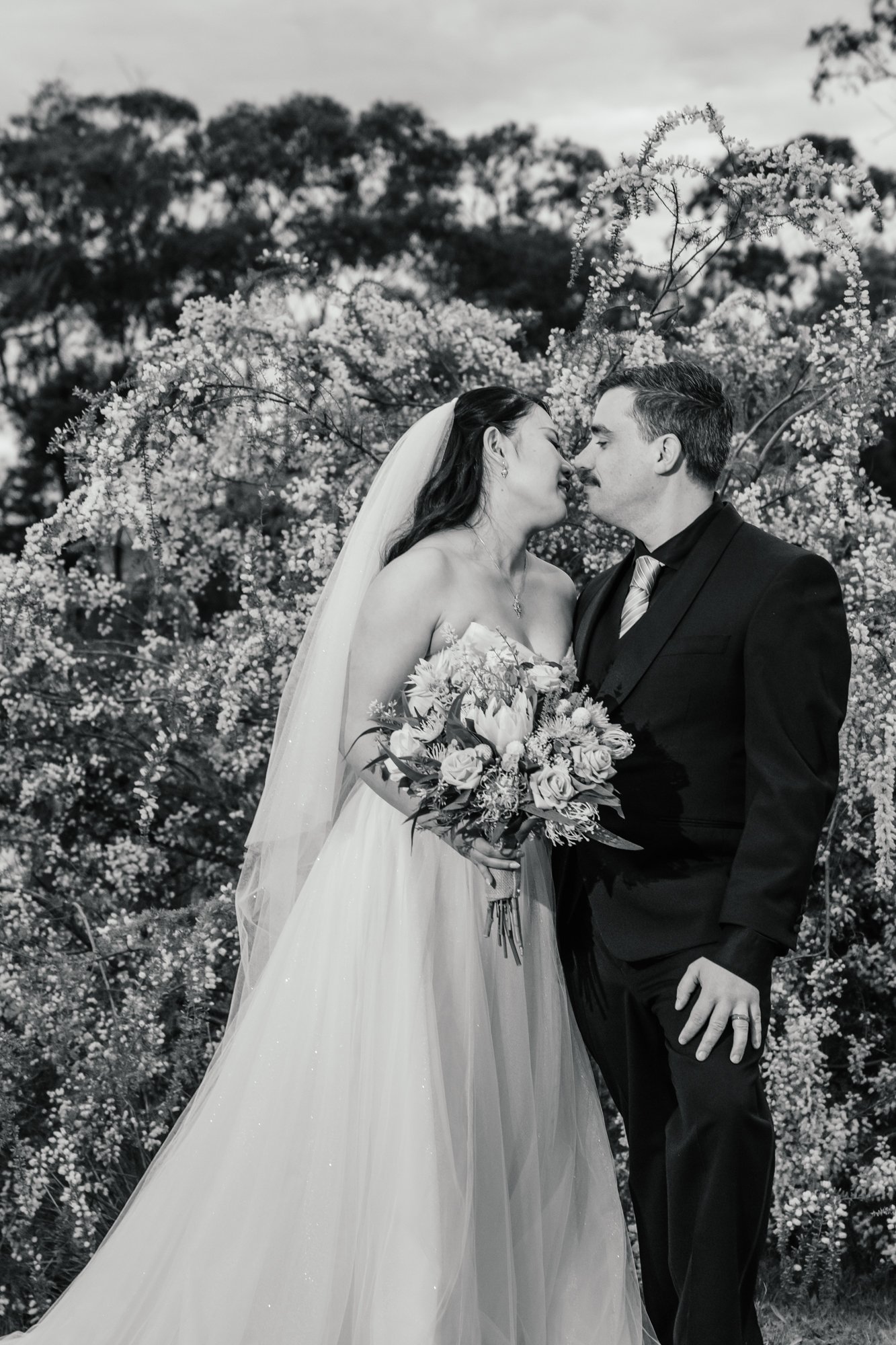 A black and white photograph of a bride and groom standing close, with their foreheads touching, surrounded by flowering trees, during a wedding celebration.