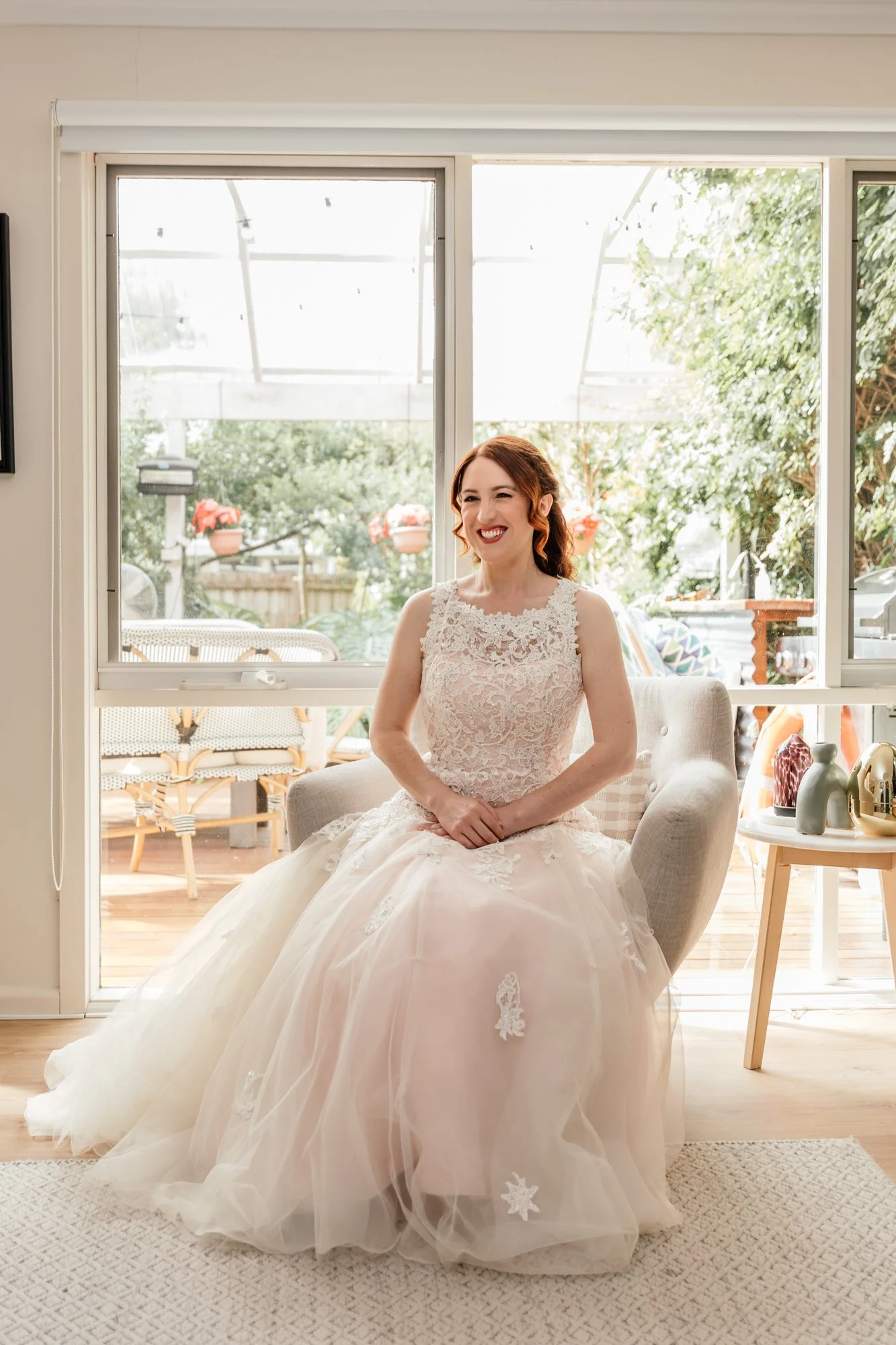 A woman in a wedding dress sitting in a living room near glass sliding doors, smiling with her eyes closed.