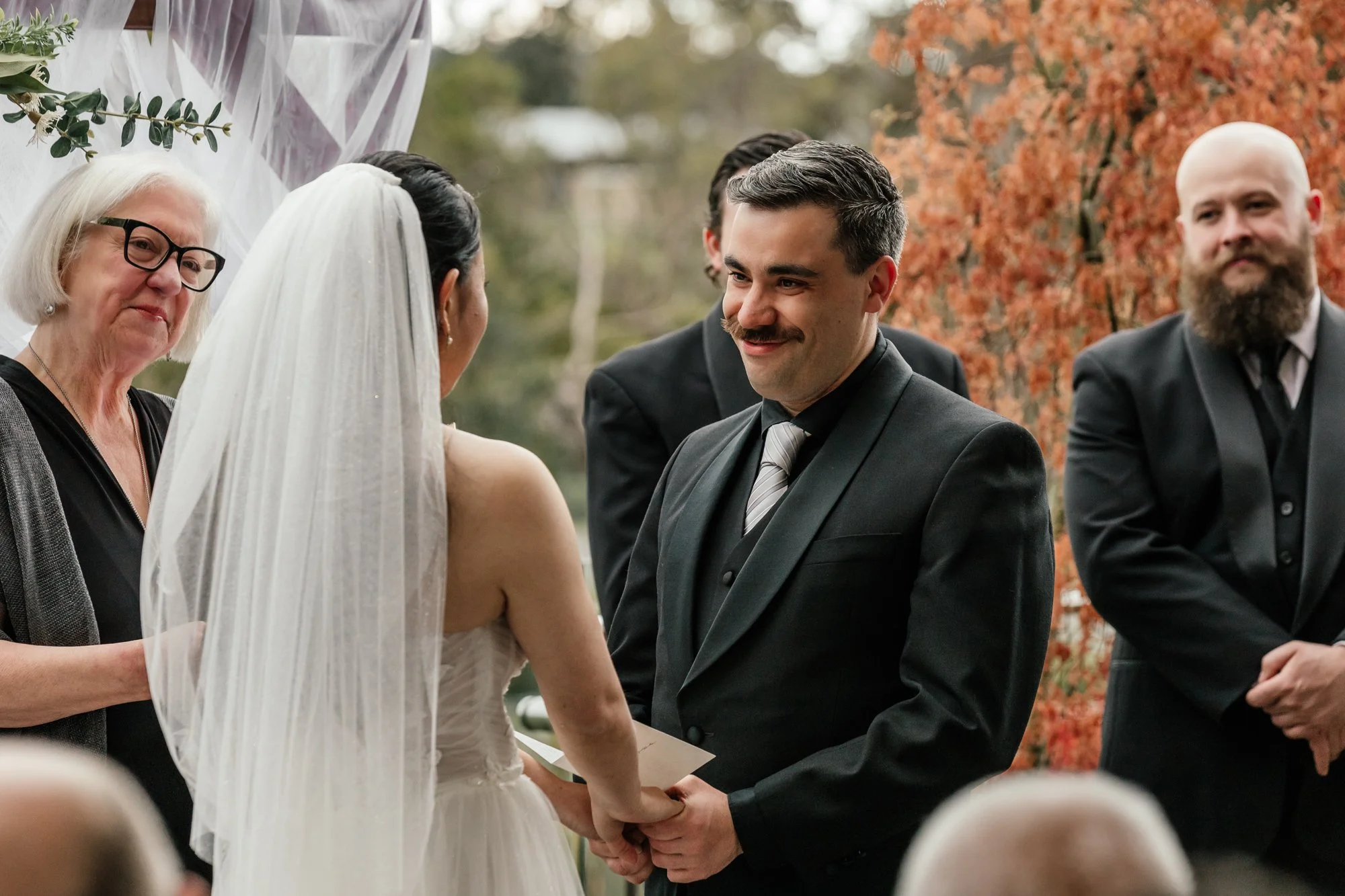 Groom and bride holding hands during wedding ceremony outdoors with officiant and groomsmen, autumn leaves background.