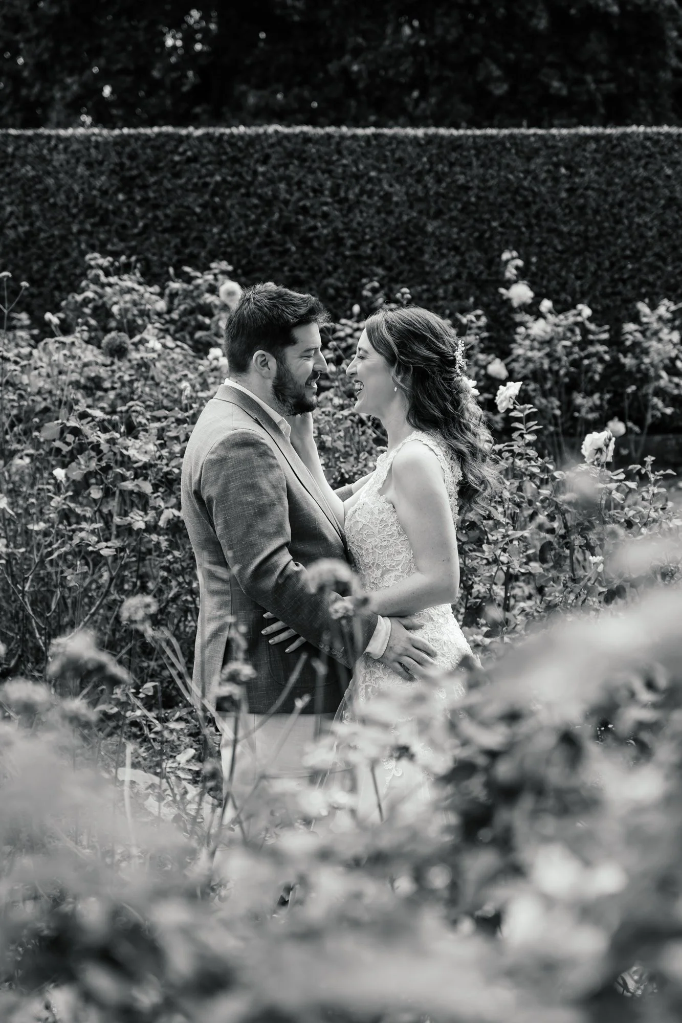 Black and white photo of a wedding couple standing close in a garden, smiling and looking into each other's eyes, surrounded by flowers and shrubbery.