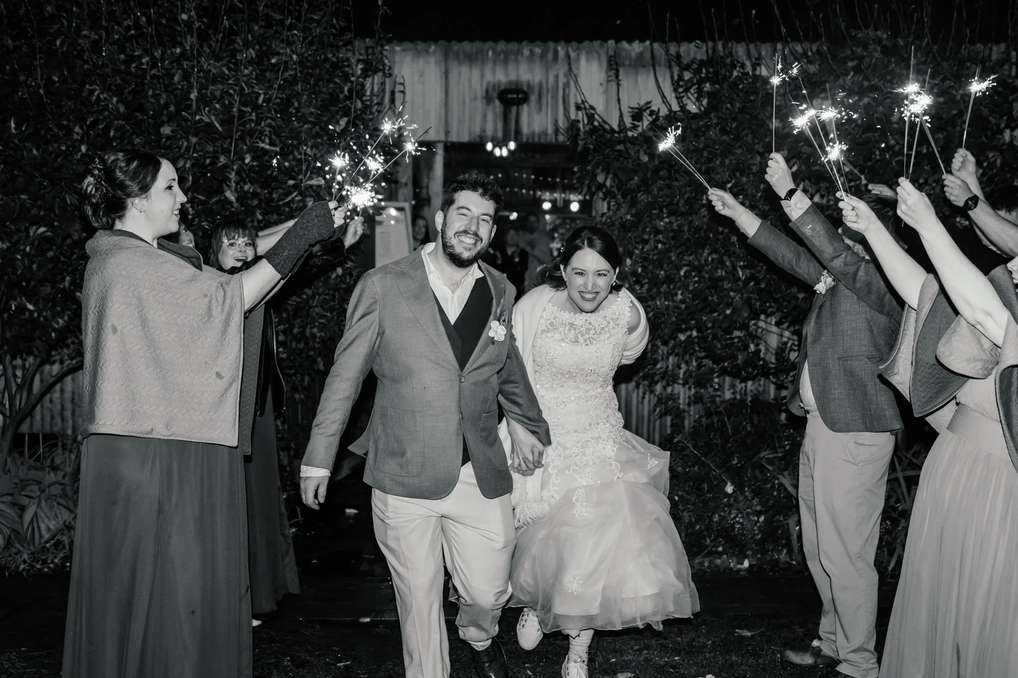 A black and white photo of a newlywed couple walking hand in hand through a celebration with guests holding sparklers on either side. The couple is smiling, with the bride wearing a wedding dress and the groom in a suit.