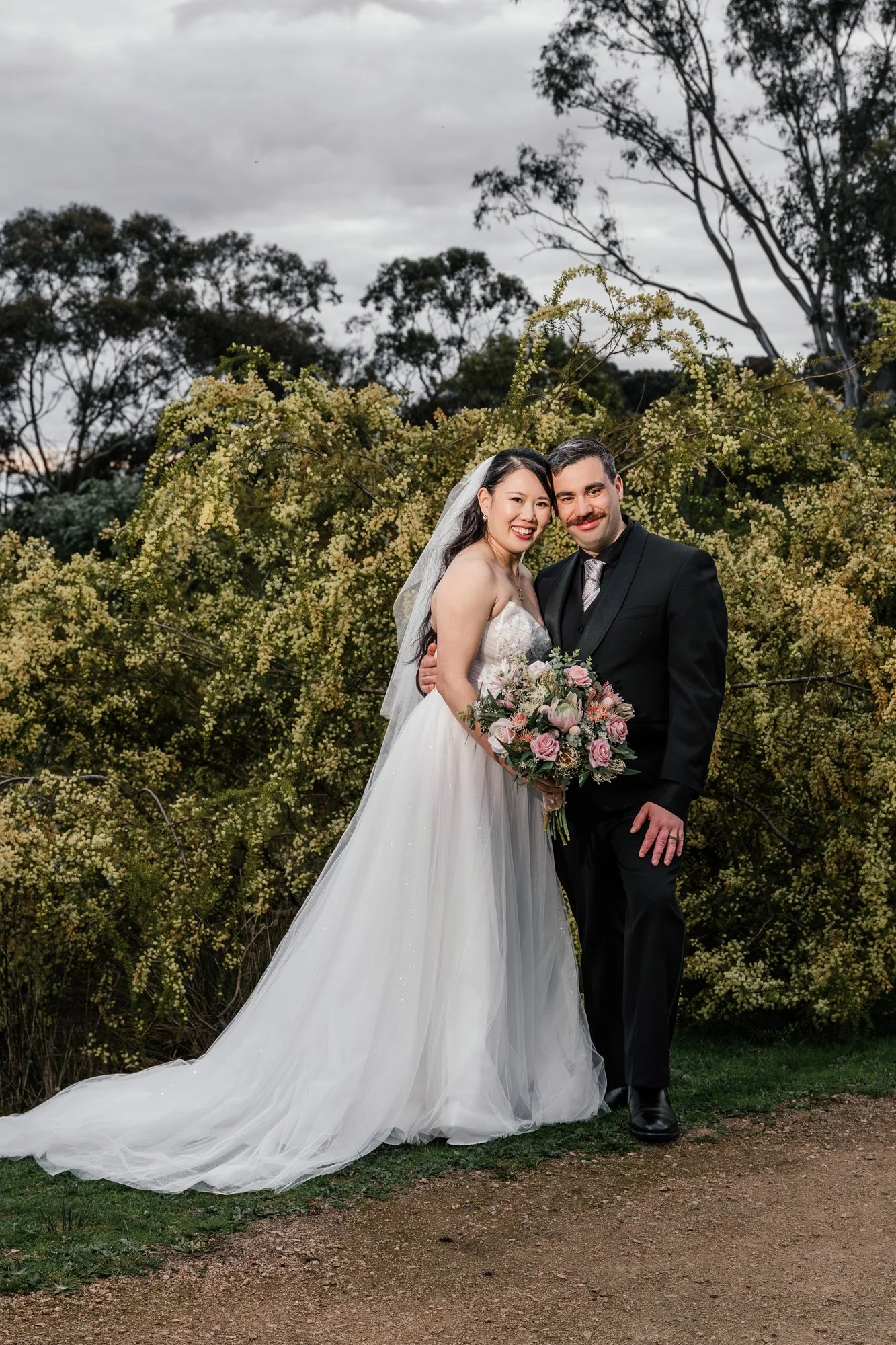 A newlywed couple poses outdoors with trees and cloudy sky in the background. The bride wears a white wedding gown with a long train and holds a bouquet of pink and white flowers. The groom wears a black suit with a striped tie. They are smiling and 