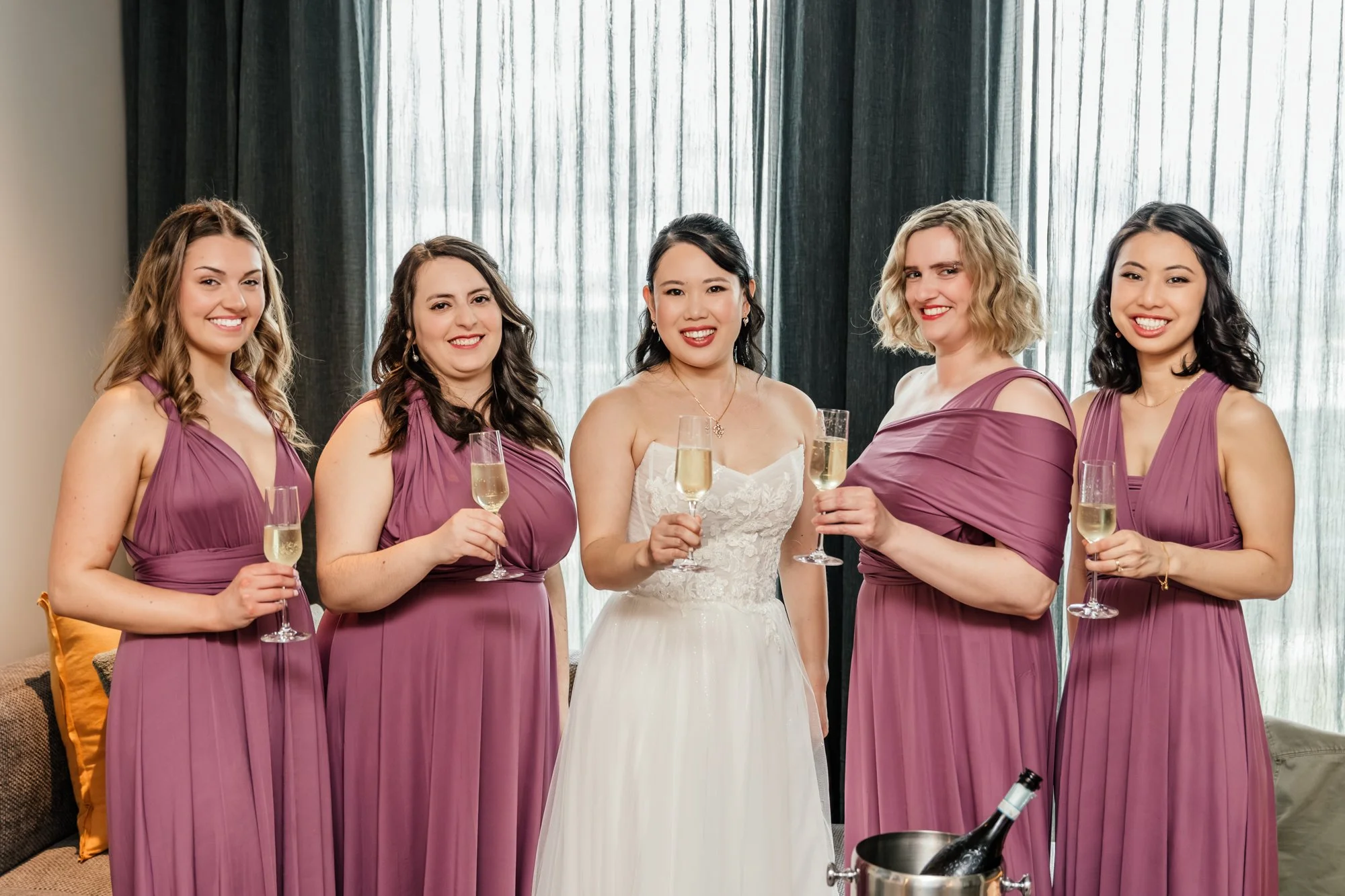 Group of five women in a celebration, with one in a white wedding dress and four in purple bridesmaid dresses, holding champagne glasses, standing in a room with large curtains and a table with a wine bucket in the foreground.