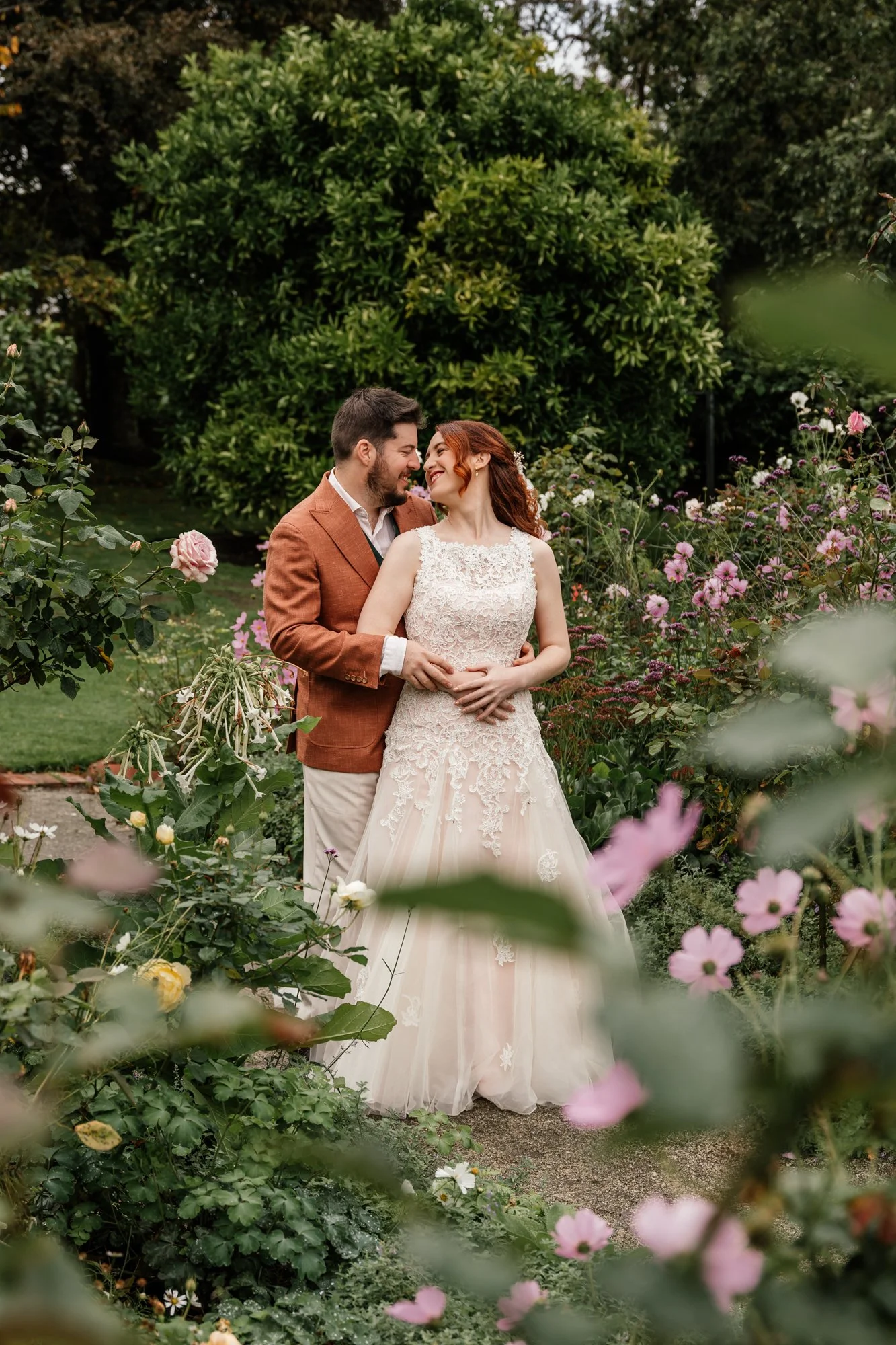 A smiling bride and groom share a close moment in a garden filled with pink and white flowers, with lush green trees in the background.
