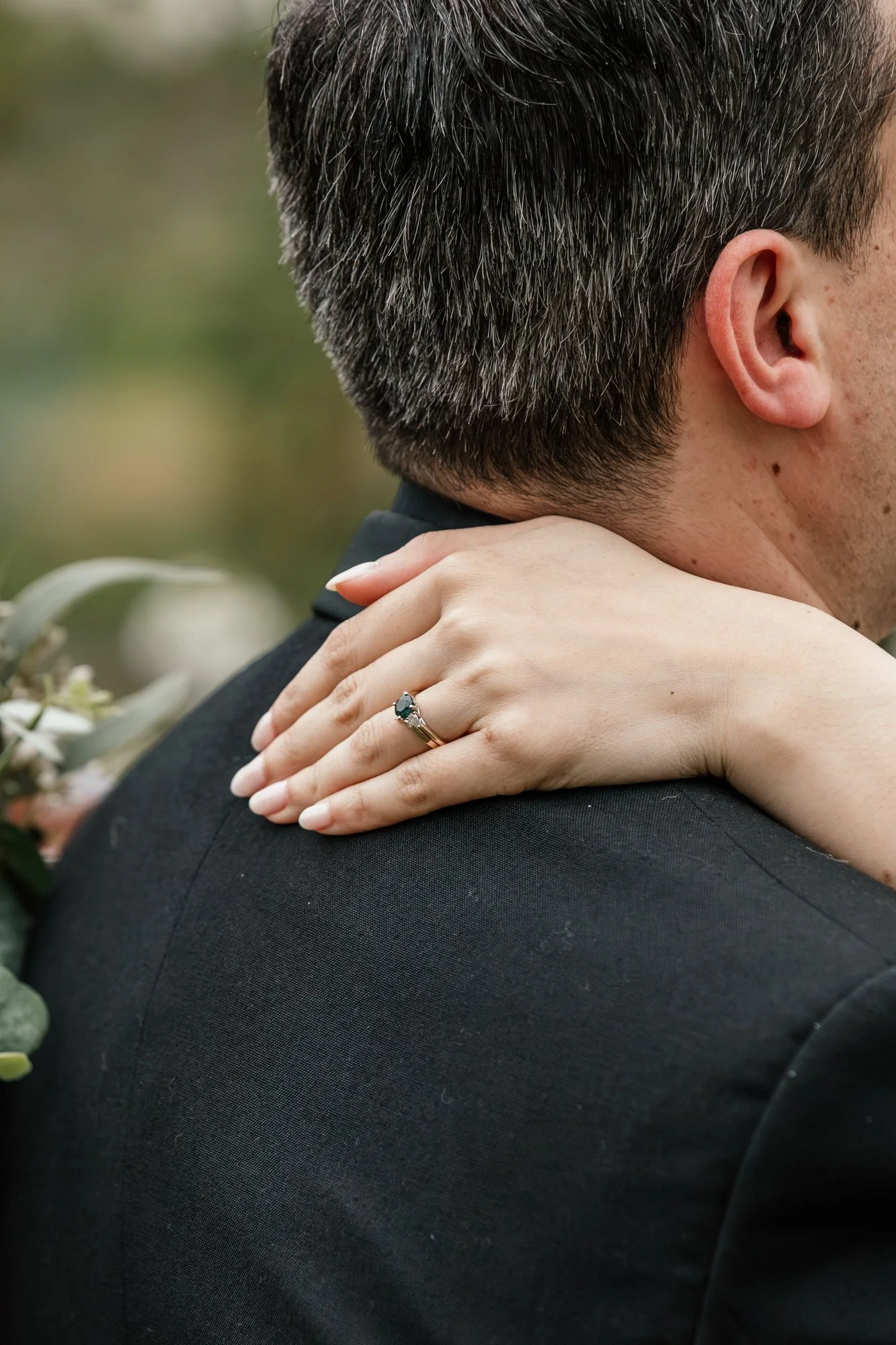 Close-up of a couple embracing, with the woman resting her hand gently on the man's shoulder, showing a ring with a large dark green gemstone. The man has short, dark hair with some gray, and is wearing a dark suit. The background is blurred with sof