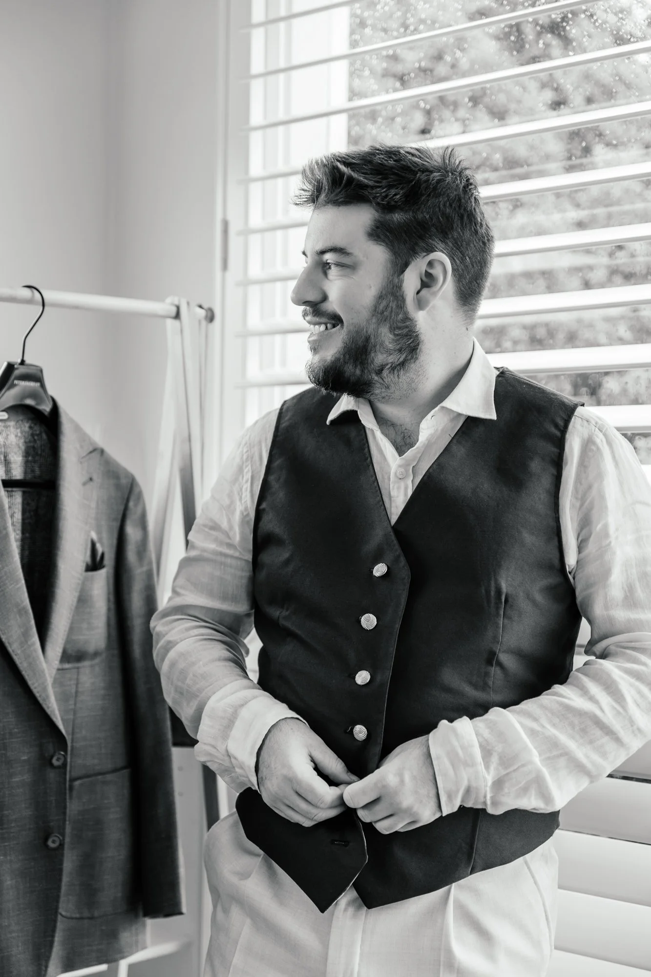 A smiling man with a beard and short hair in a vest, sitting near a window with blind slats, next to a clothing rack with a suit jacket. The photo is in black and white.