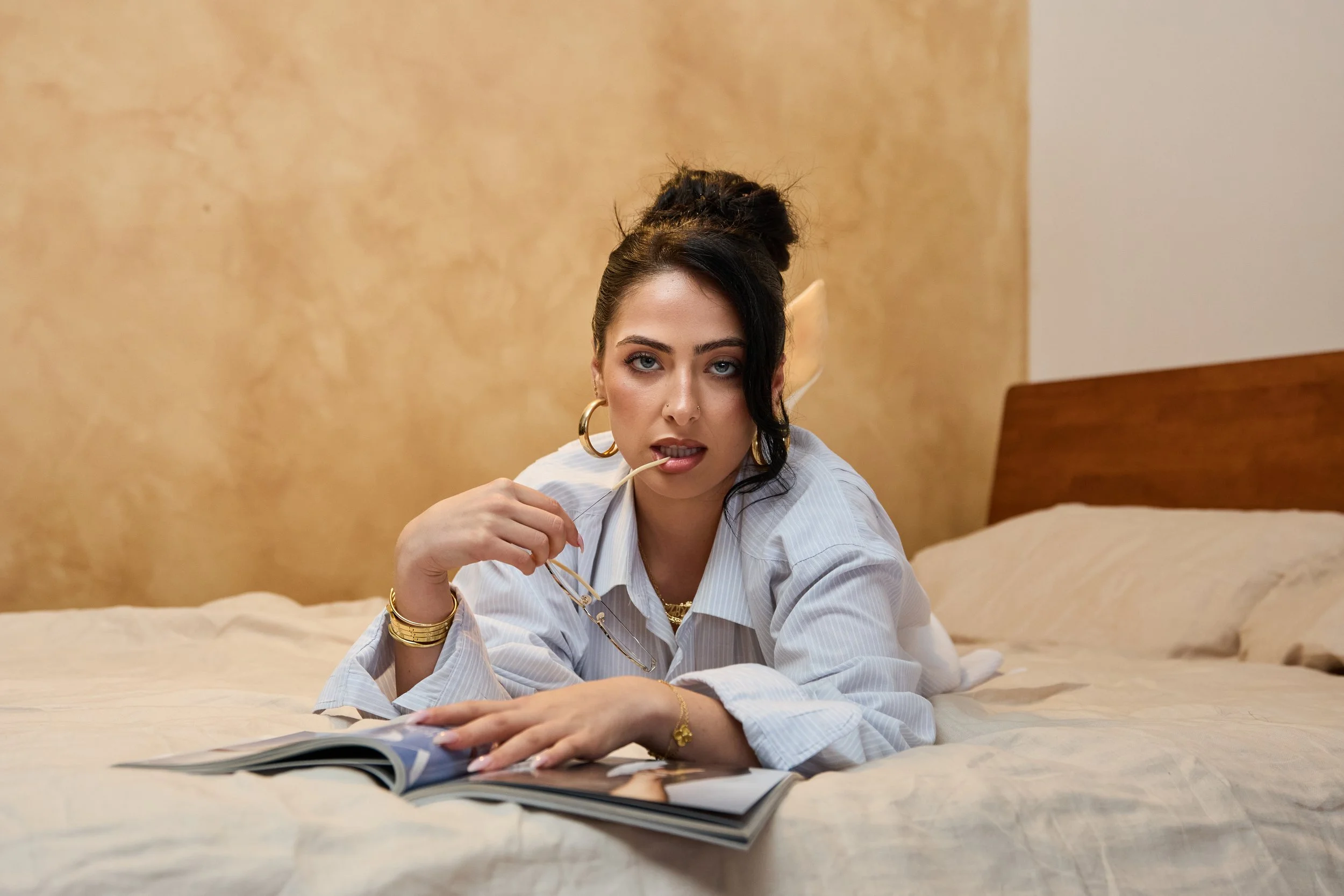 A woman with dark hair styled in an updo, wearing large hoop earrings, gold jewelry, and a striped shirt, lying on a bed and reading a magazine, holding a pair of glasses in her mouth, with a yellow textured wall and a wooden headboard in the backgro