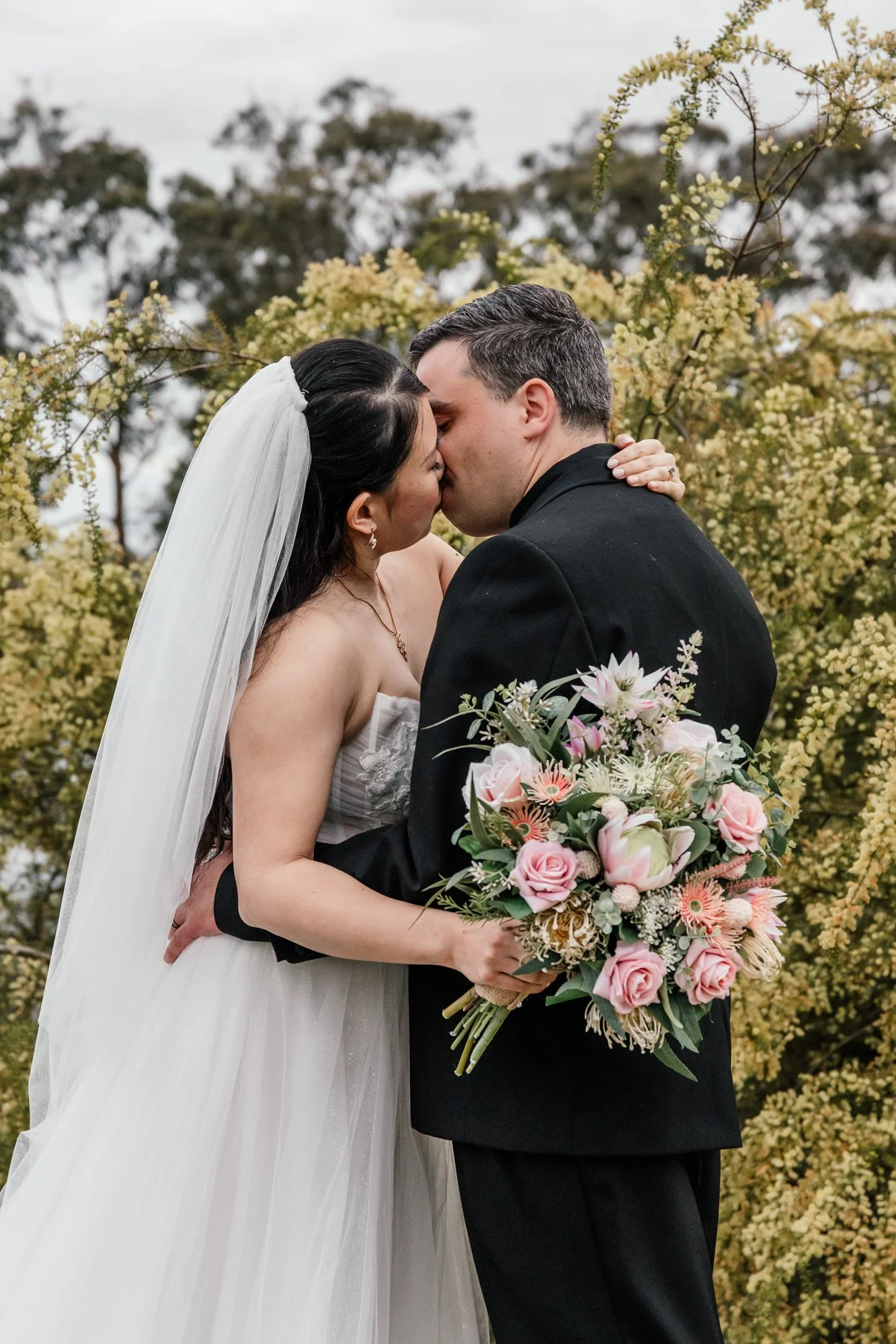 A bride and groom kissing outdoors with flowering bushes in the background. The bride is wearing a white wedding dress and veil, holding a bouquet of pink and cream flowers. The groom is dressed in a black suit and top shirt.
