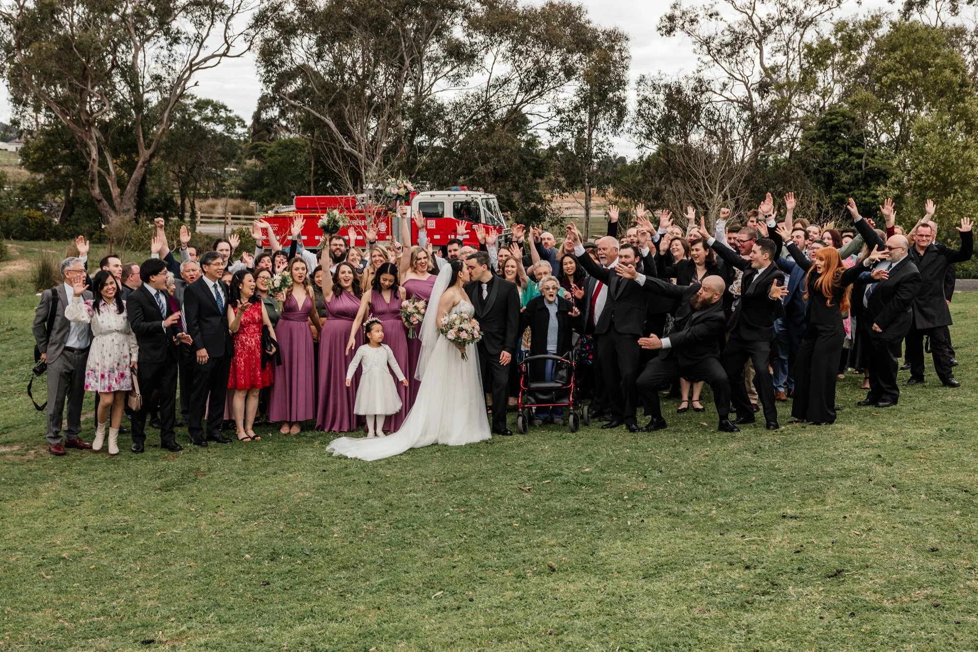 A large group of people gathered outdoors for a wedding celebration, with a bride and groom in the center, flowers, and a fire truck decorated with flowers in the background.