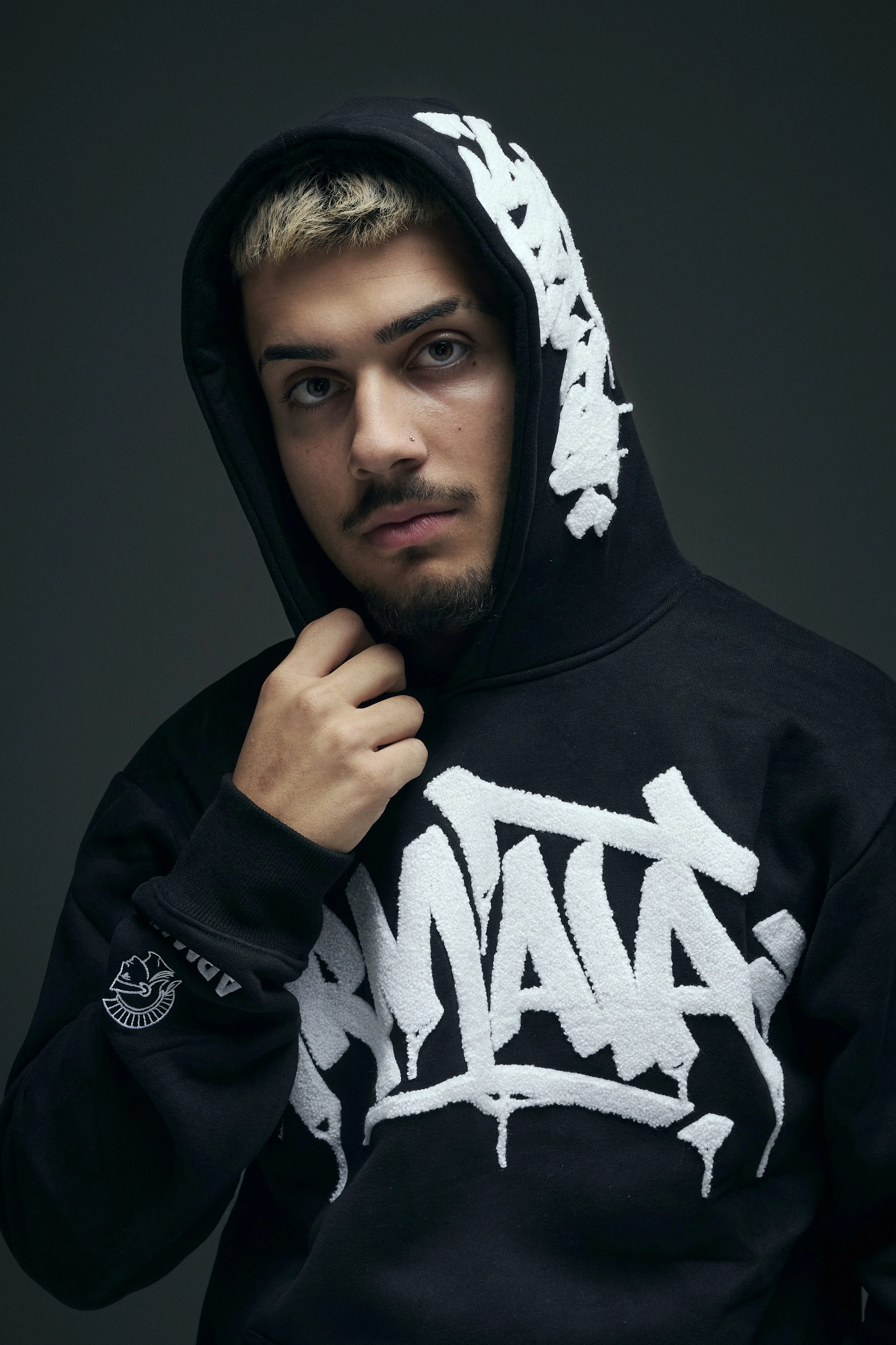 A young man with blonde hair, facial hair, and blue eyes wearing a black hoodie with white text and designs, pulling the hoodie slightly to reveal part of his face, against a dark gray background.