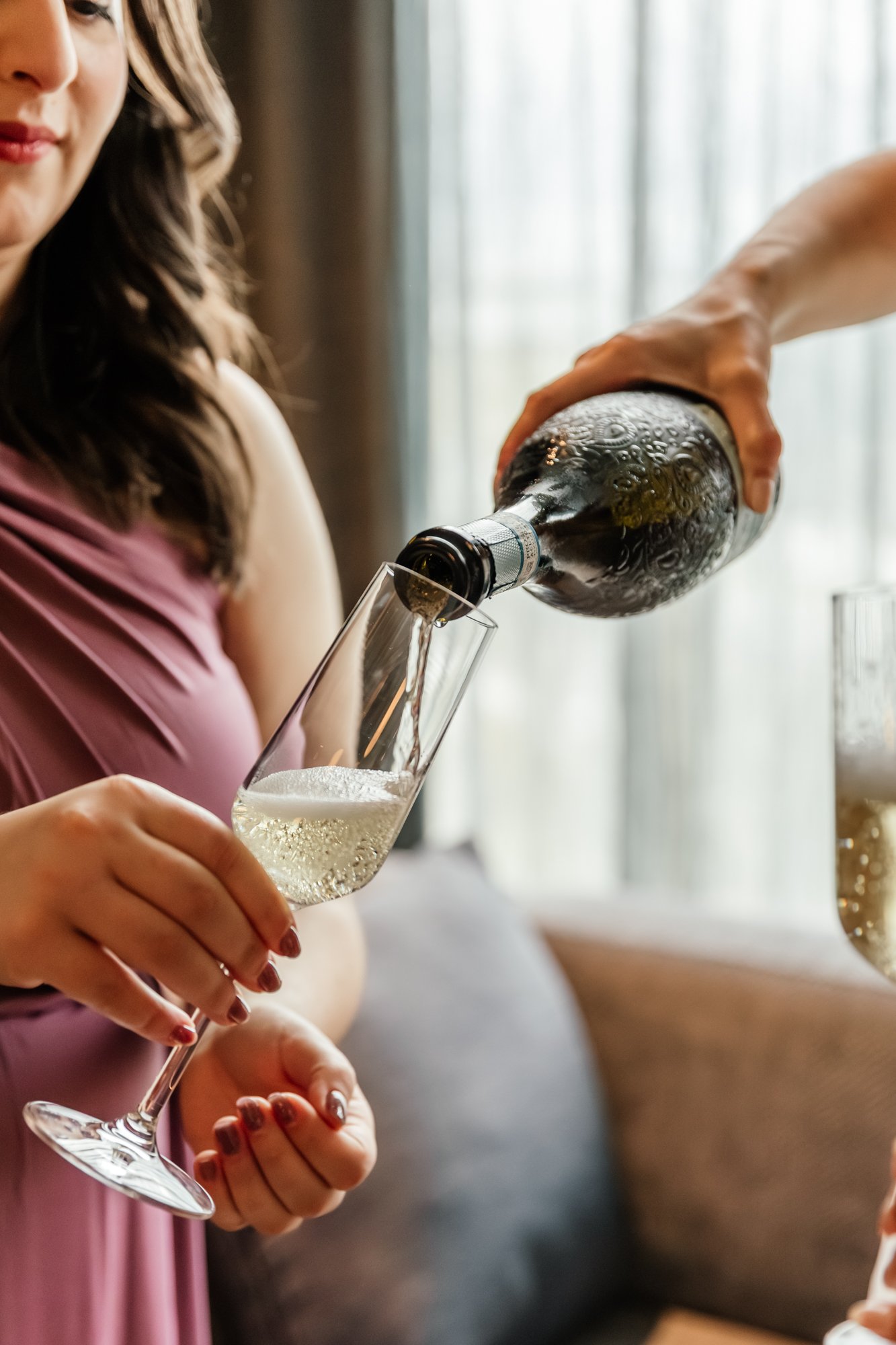A woman in a pink dress is pouring champagne into a glass, with her nails painted red, in front of a bright window with curtains.