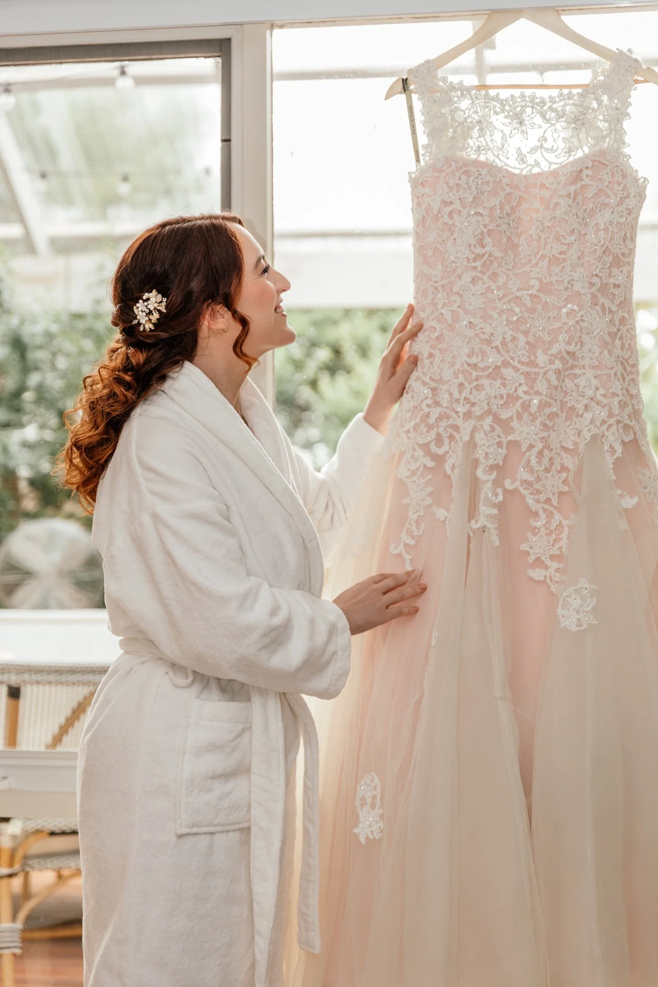 A woman in a white bathrobe smiling and looking at a pink lace wedding dress on a hanger.