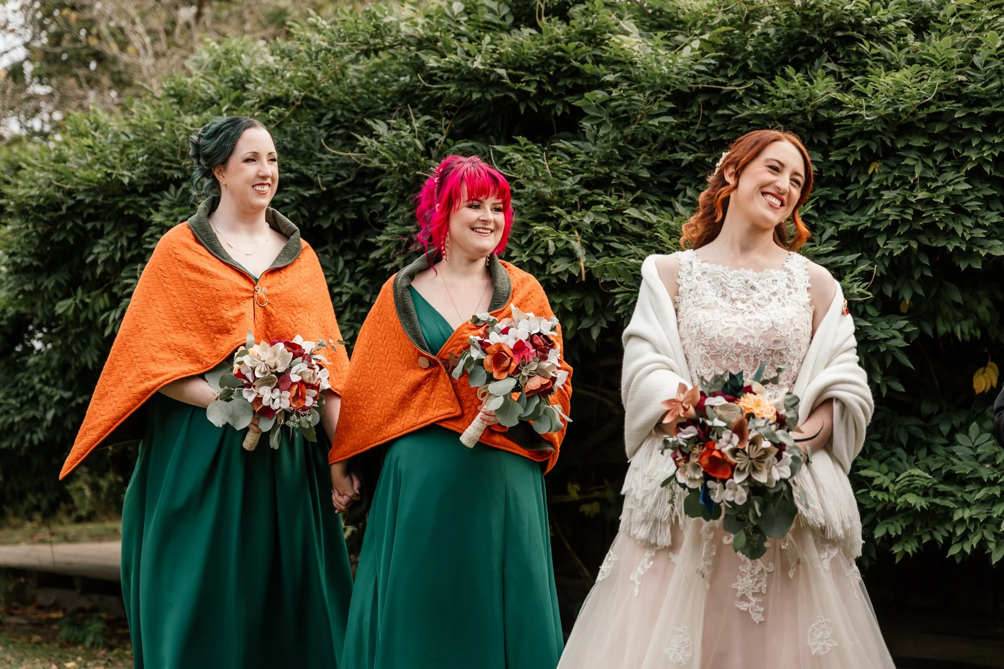 Three women stand outdoors in front of green foliage. They are dressed formally, with two women wearing matching green dresses and orange capes, and the third woman wearing a white lace wedding gown. They are smiling and holding bouquets of flowers.
