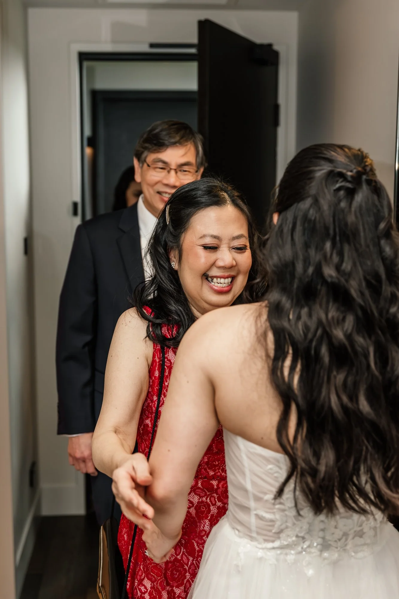 People at a wedding or formal event, with a woman in a red dress hugging another woman in a white dress, and a man in a suit smiling in the background.