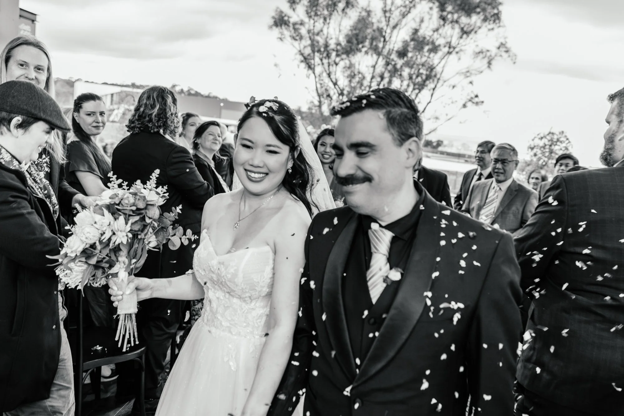 A wedding celebration with a bride in a white wedding dress and a groom in a dark suit, walking hand in hand while being showered with confetti, surrounded by friends and family outdoors.