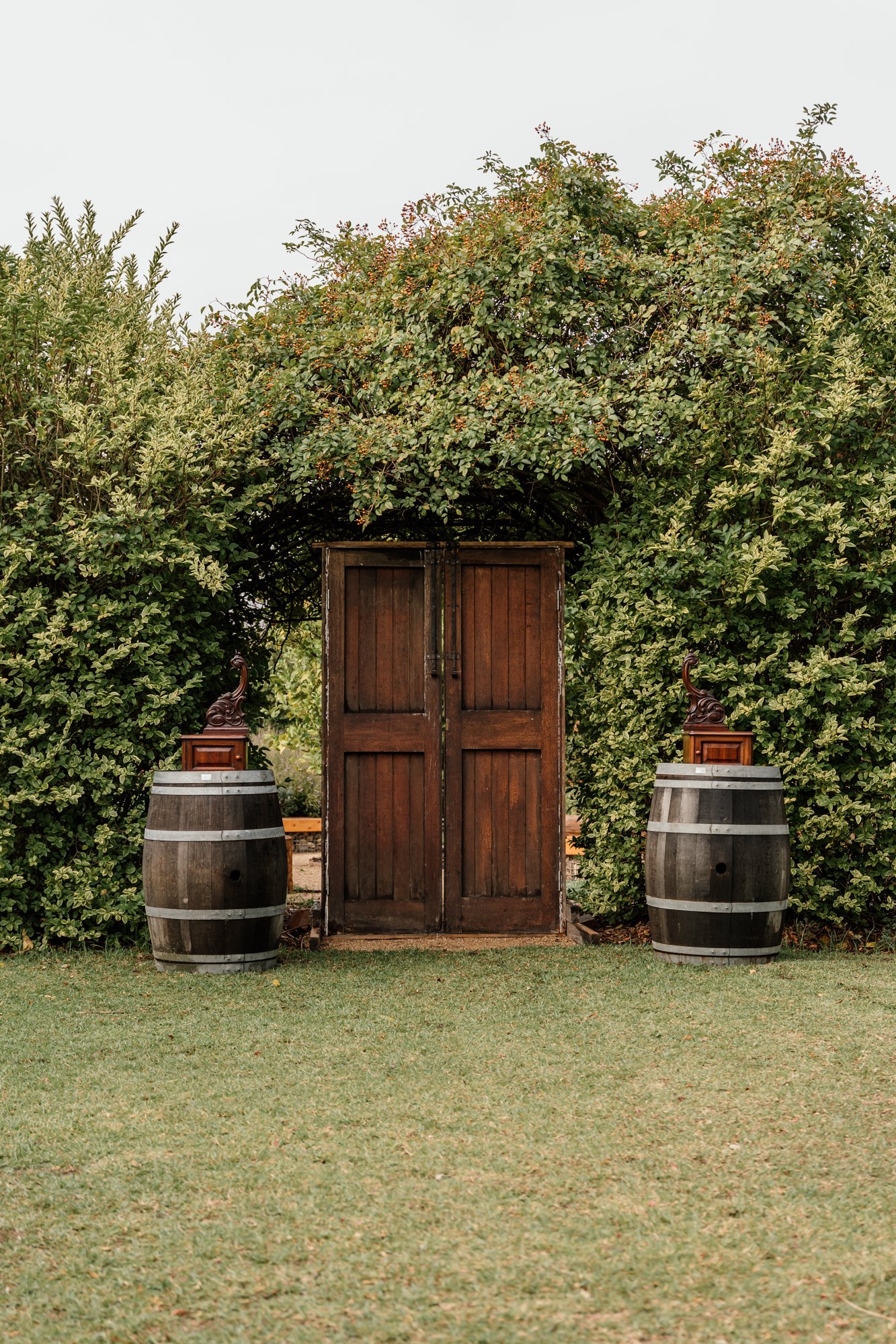 A rustic outdoor scene with a wooden gate framed by lush green bushes and trees, flanked by two wine barrels with decorative carved wood accents on top.