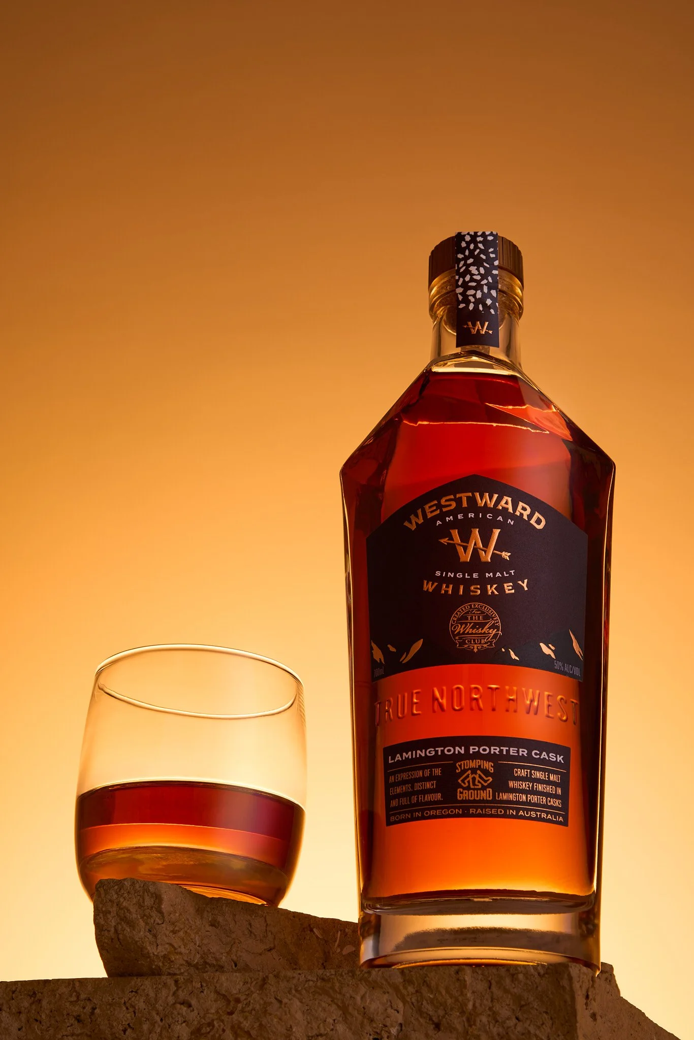 A bottle of Westward American single malt whiskey with a glass of whiskey beside it, set against a warm sunset background.