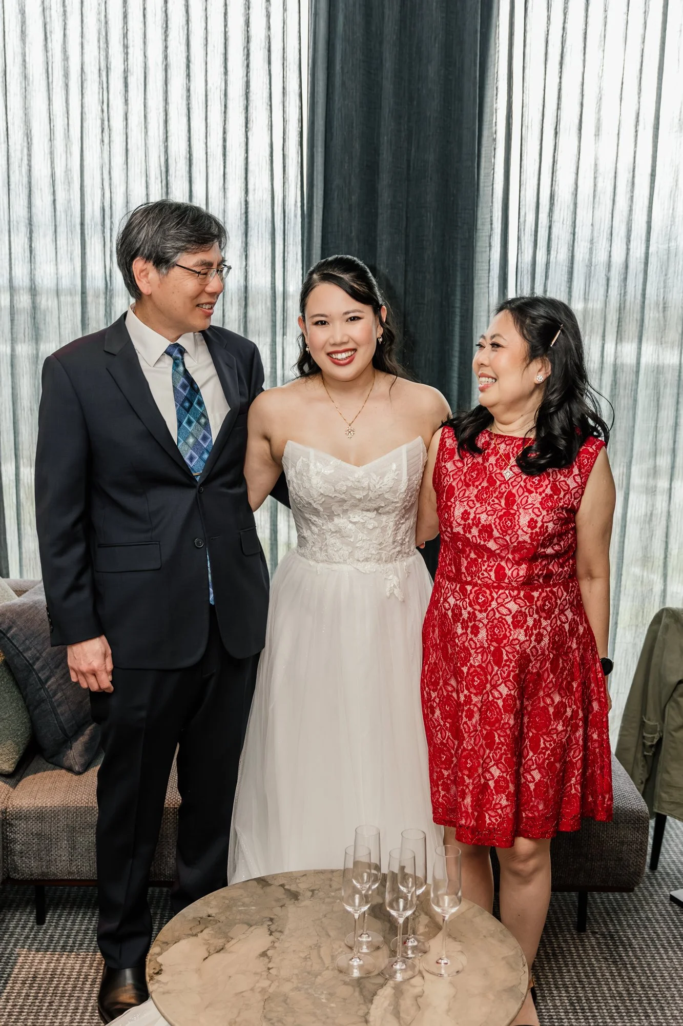 A group of three people, a man in a dark suit and glasses, a woman in a white dress, and another woman in a red lace dress, standing together and smiling in a stylish indoor setting with a marble table holding several empty champagne glasses in front