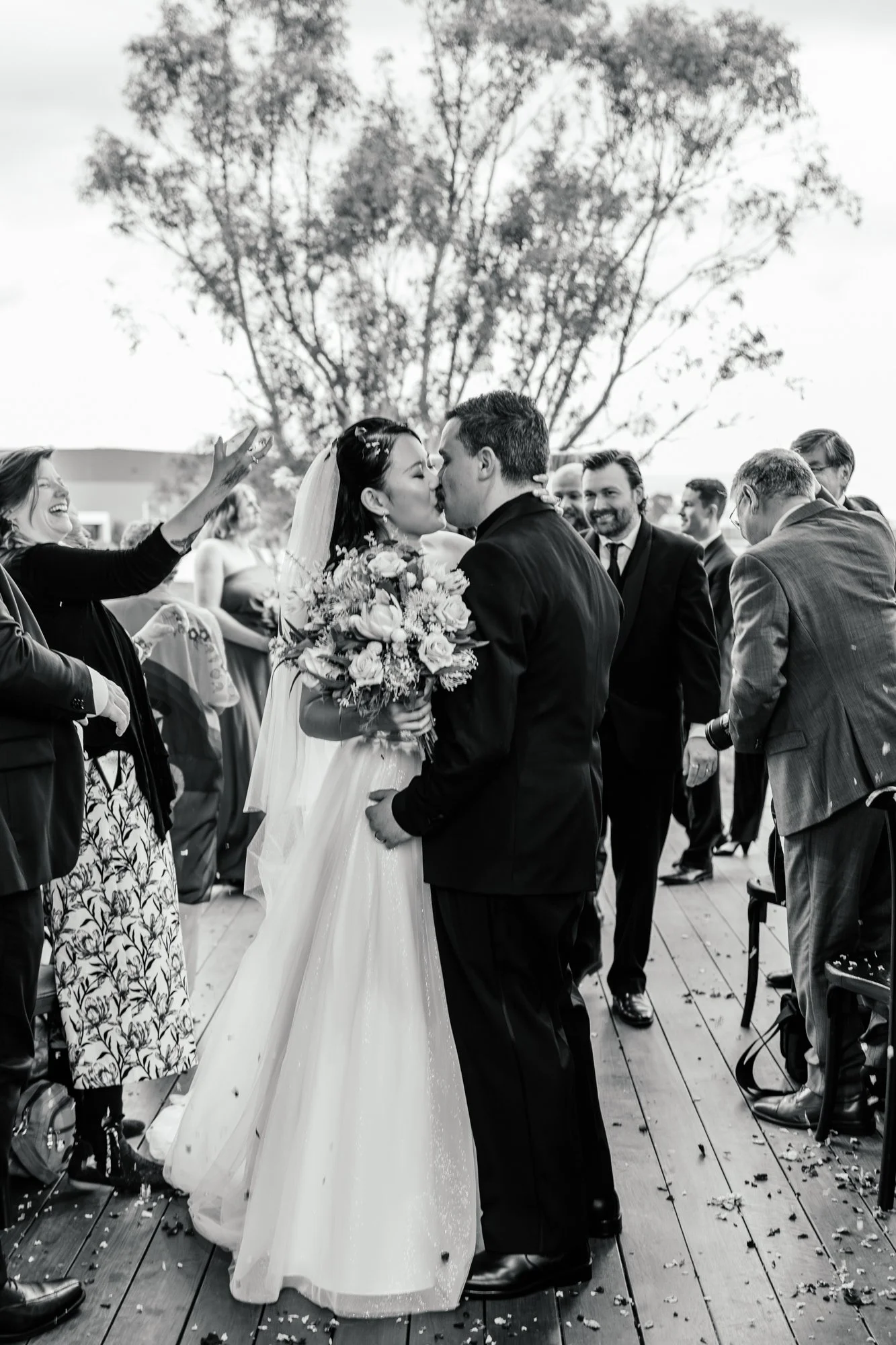 A black-and-white photo of a wedding scene with a couple kissing. The bride holds a large bouquet of flowers, and guests surround them celebrating. There is a big tree in the background and confetti on the wooden floor.