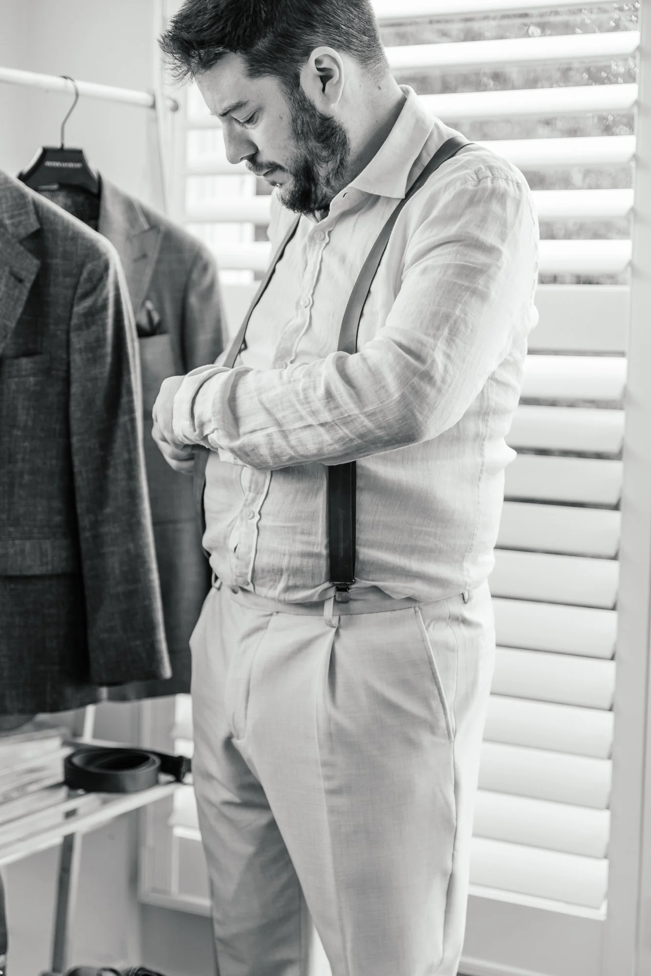A man with a beard and mustache, wearing a light-colored dress shirt with suspenders and light-colored trousers, is standing indoors near a window with plantation shutters. The man appears to be adjusting his cuff or sleeve and is looking down. To th