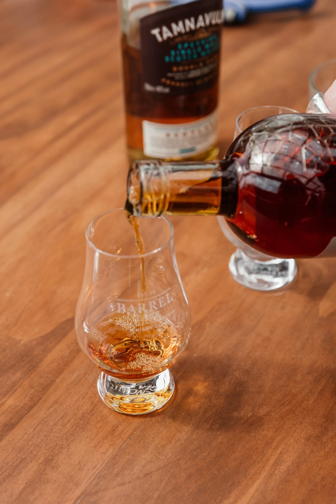 A glass with amber-colored whiskey being poured from a bottle with a black label, and another bottle of whiskey in the background on a wooden surface.