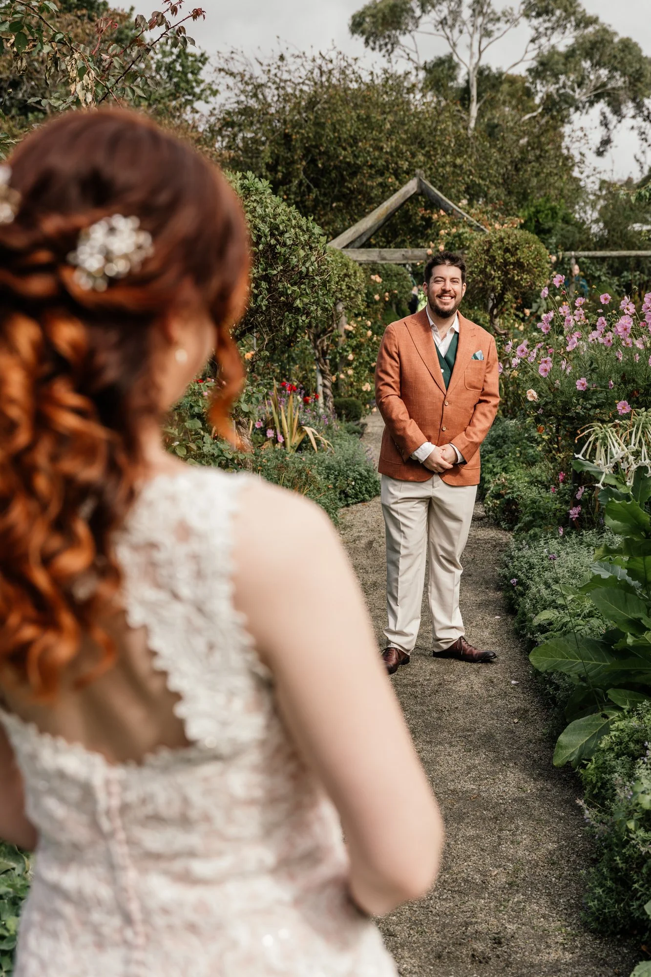 A man in a brown blazer and beige pants smiling at a woman with red hair in a garden with colorful flowers.