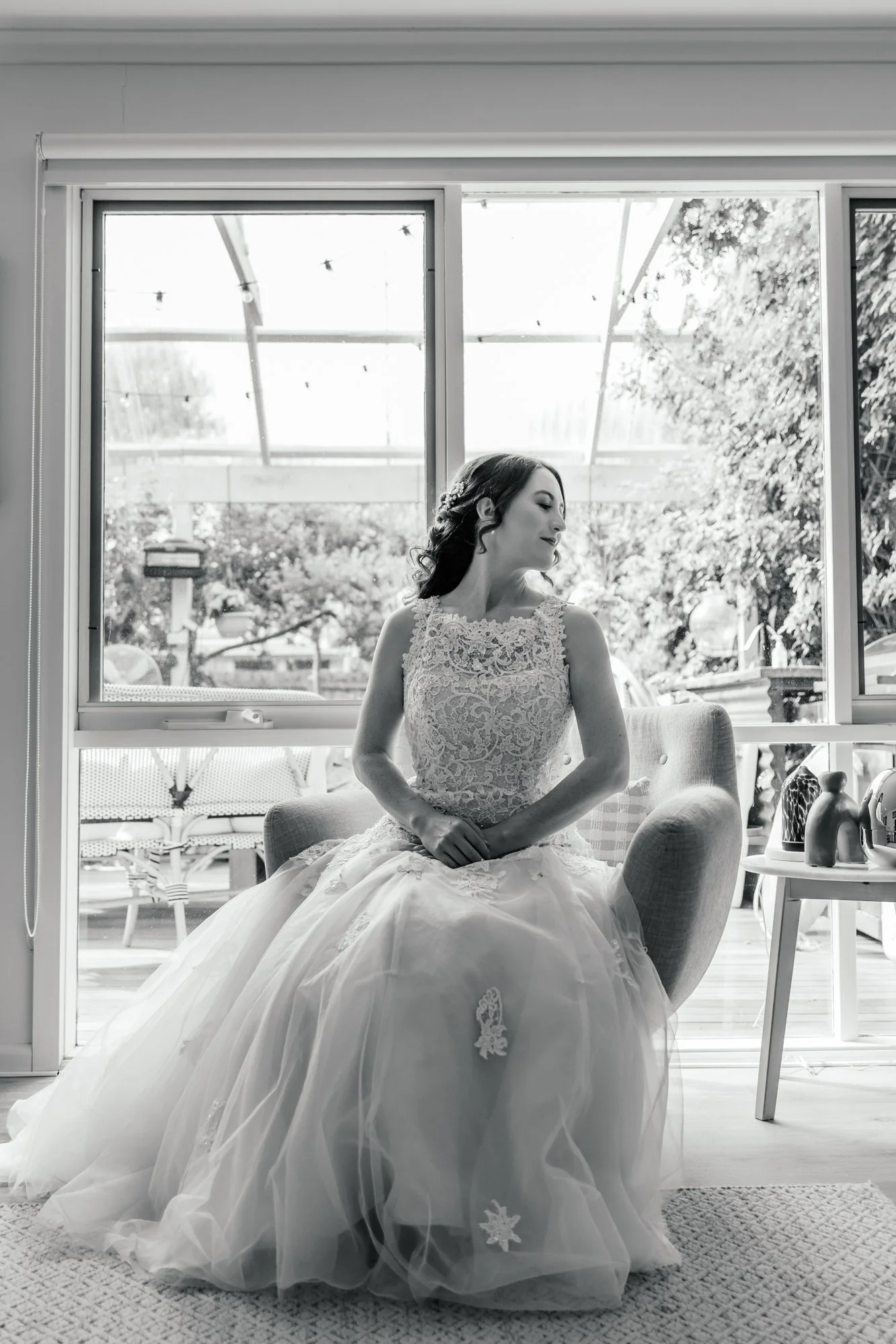 Black and white photo of a woman in a wedding dress sitting on a chair beside a large window, with trees and outdoor furniture visible outside.