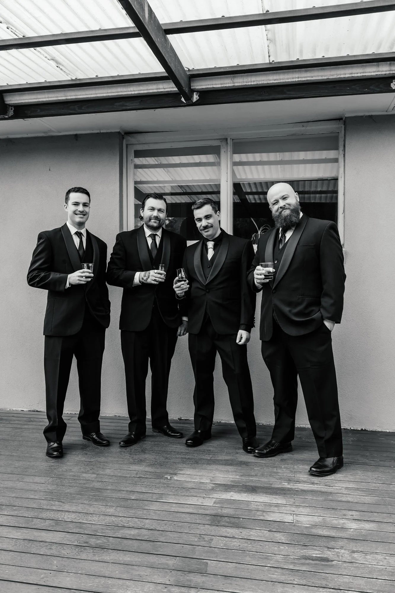 Four men in tuxedos standing outside on a wooden deck, holding drinks, in front of a wall with large windows.