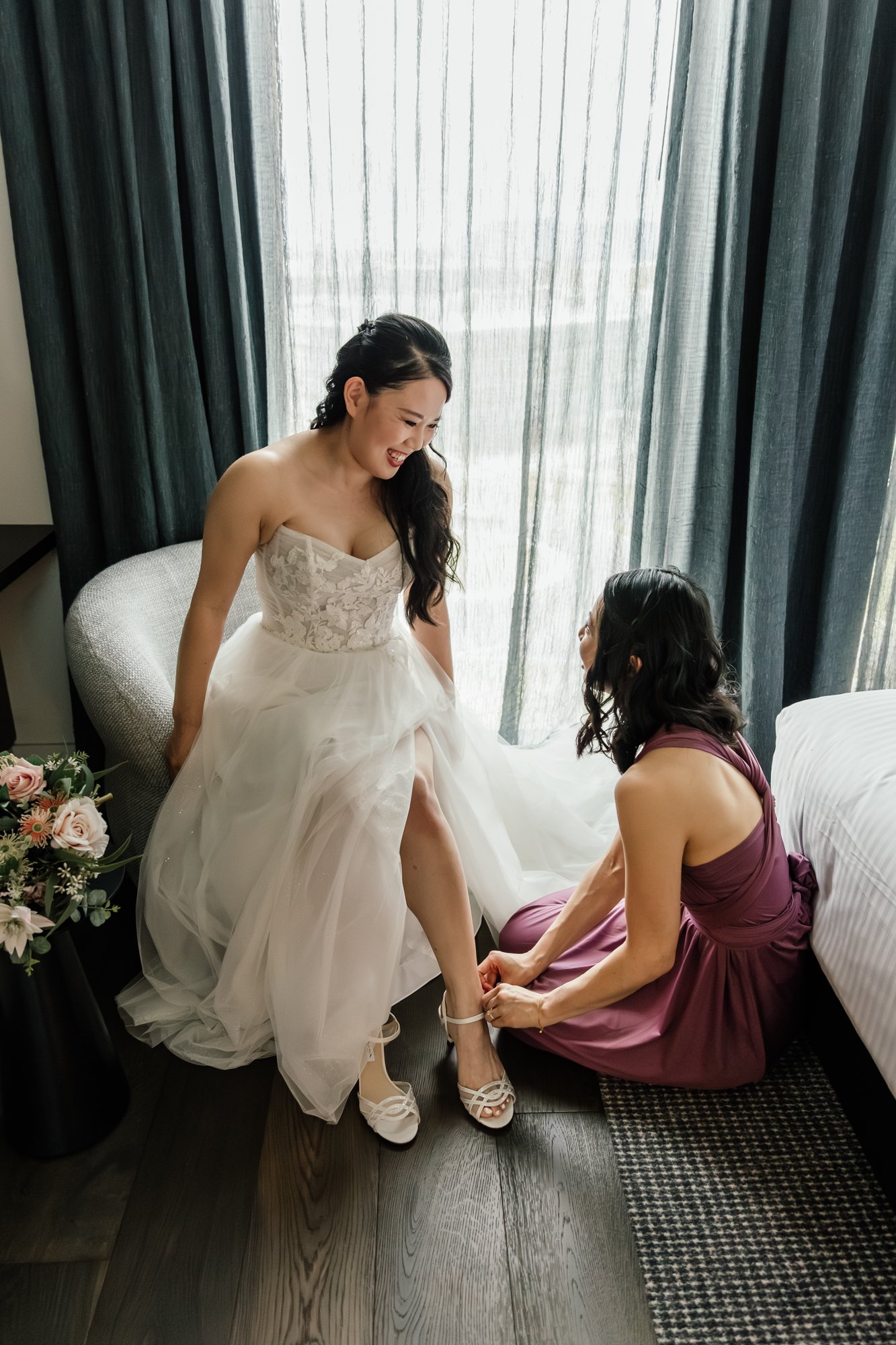 A bride in a wedding dress sitting on a chair while a woman in a purple dress helps her put on her high-heeled shoes. They are in a room with large windows and dark curtains, with a bouquet of flowers on a small table nearby.
