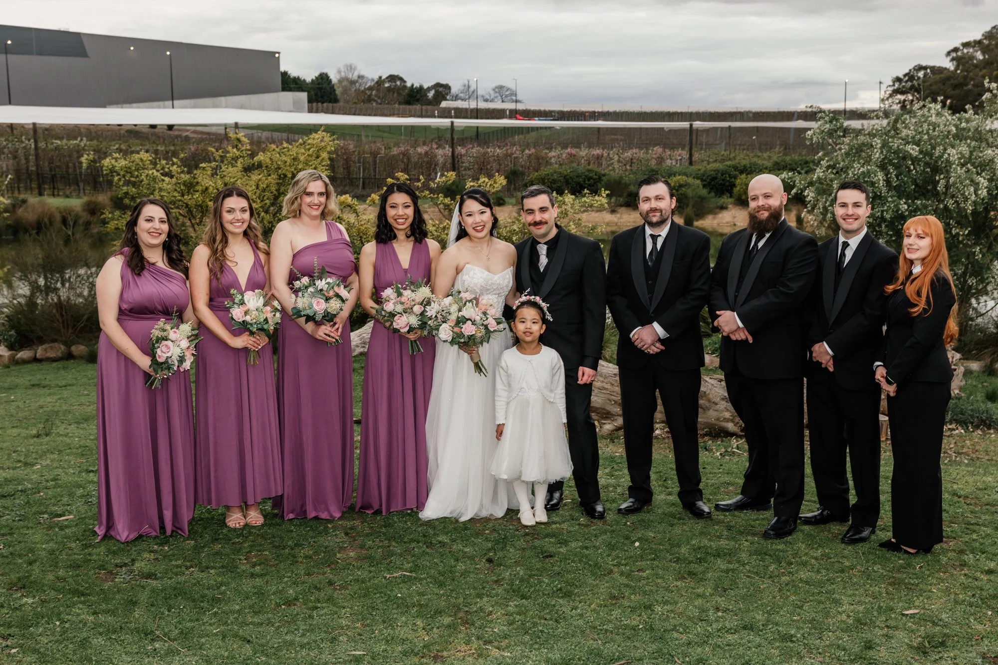 A group of wedding guests, including bridesmaids in purple dresses, a bride in a white dress, and groomsmen in black suits, standing outdoors on grass with trees and a building in the background.