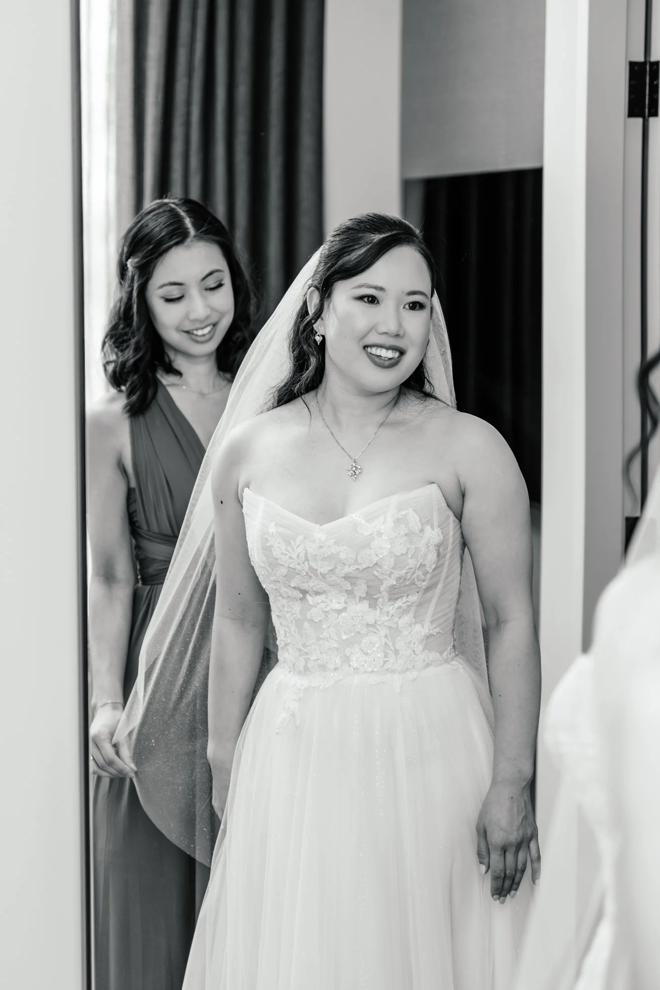 A bride in a wedding dress looking at herself in a mirror, with a bridesmaid behind her smiling and adjusting her dress. The image is in black and white.