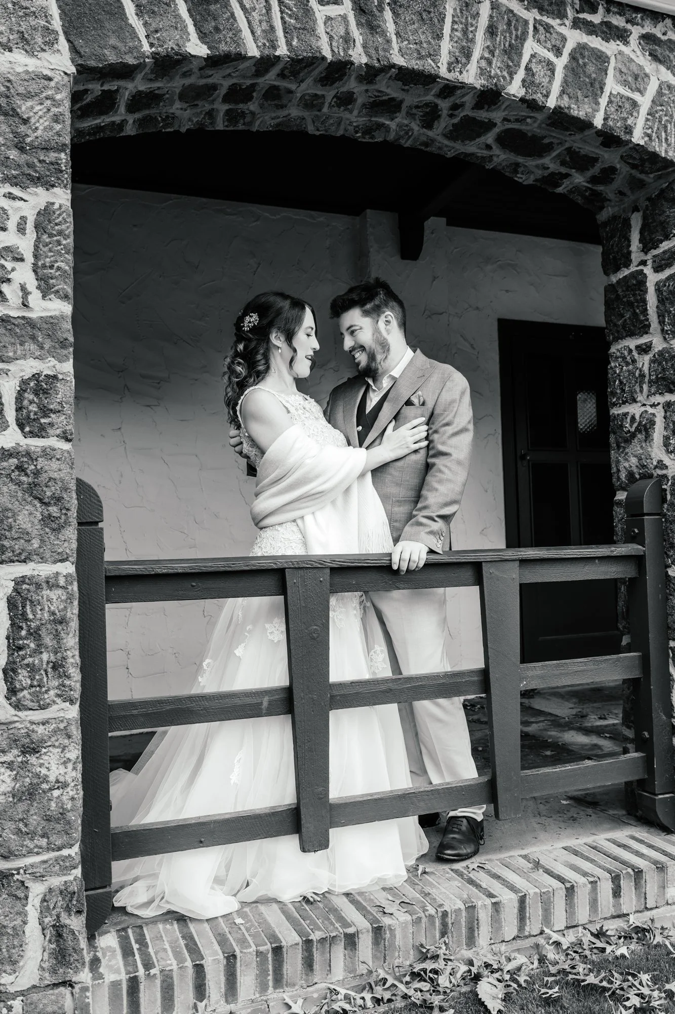 A black-and-white photo of a happy couple on their wedding day standing by a stone archway. They are gazing at each other and smiling, with the woman wearing a wedding dress and the man in a suit. The scene is set outdoors near a brick building.