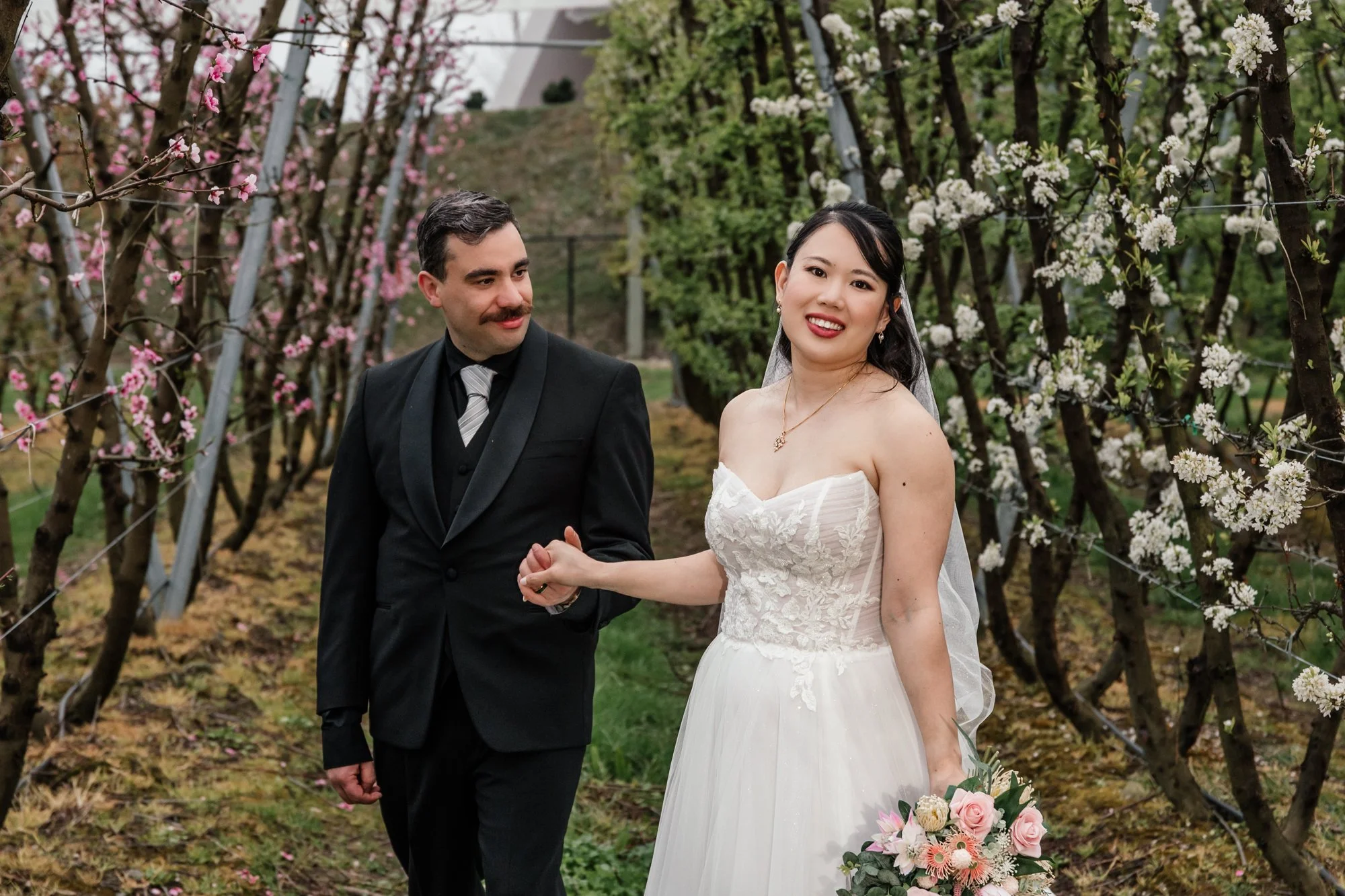 A bride and groom holding hands and smiling outdoors among blooming trees, with pink and white flowers, during a wedding ceremony.