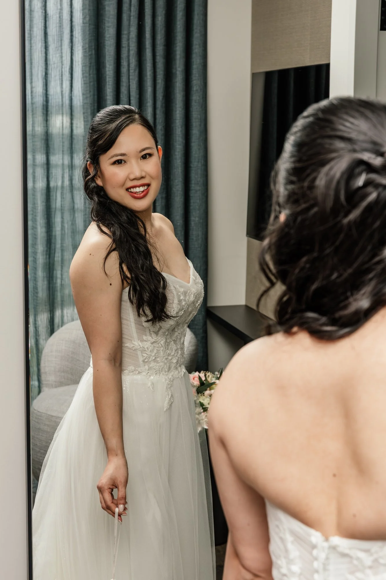 A woman in a wedding dress standing in front of a mirror, smiling, with another woman in a dress with her back to the camera, in a room with curtains and a chair.