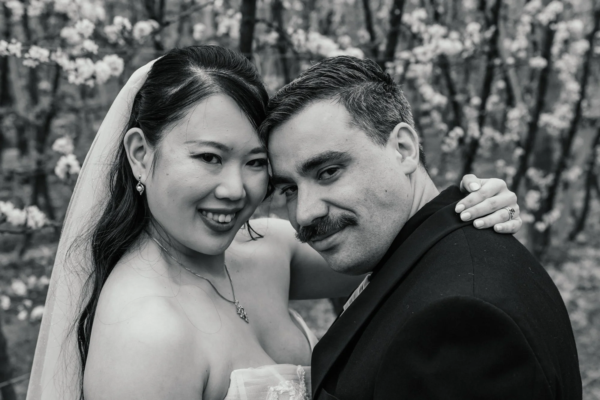 Black and white photo of a smiling woman in wedding dress and groom posing closely outdoors, with trees and flowers in the background.