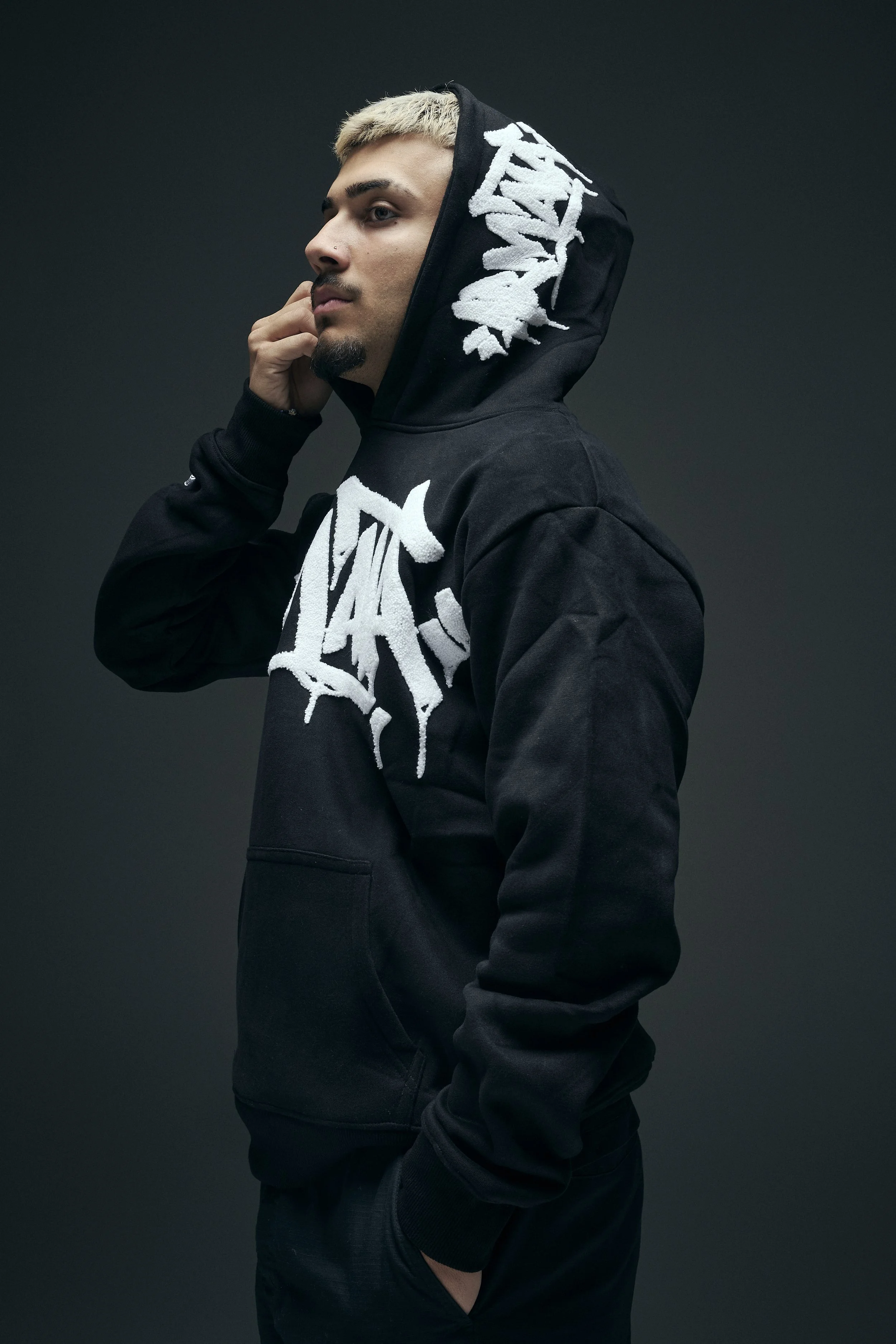 A young man with short blonde hair and facial hair wearing a black hoodie with white graphic text, standing against a dark background.