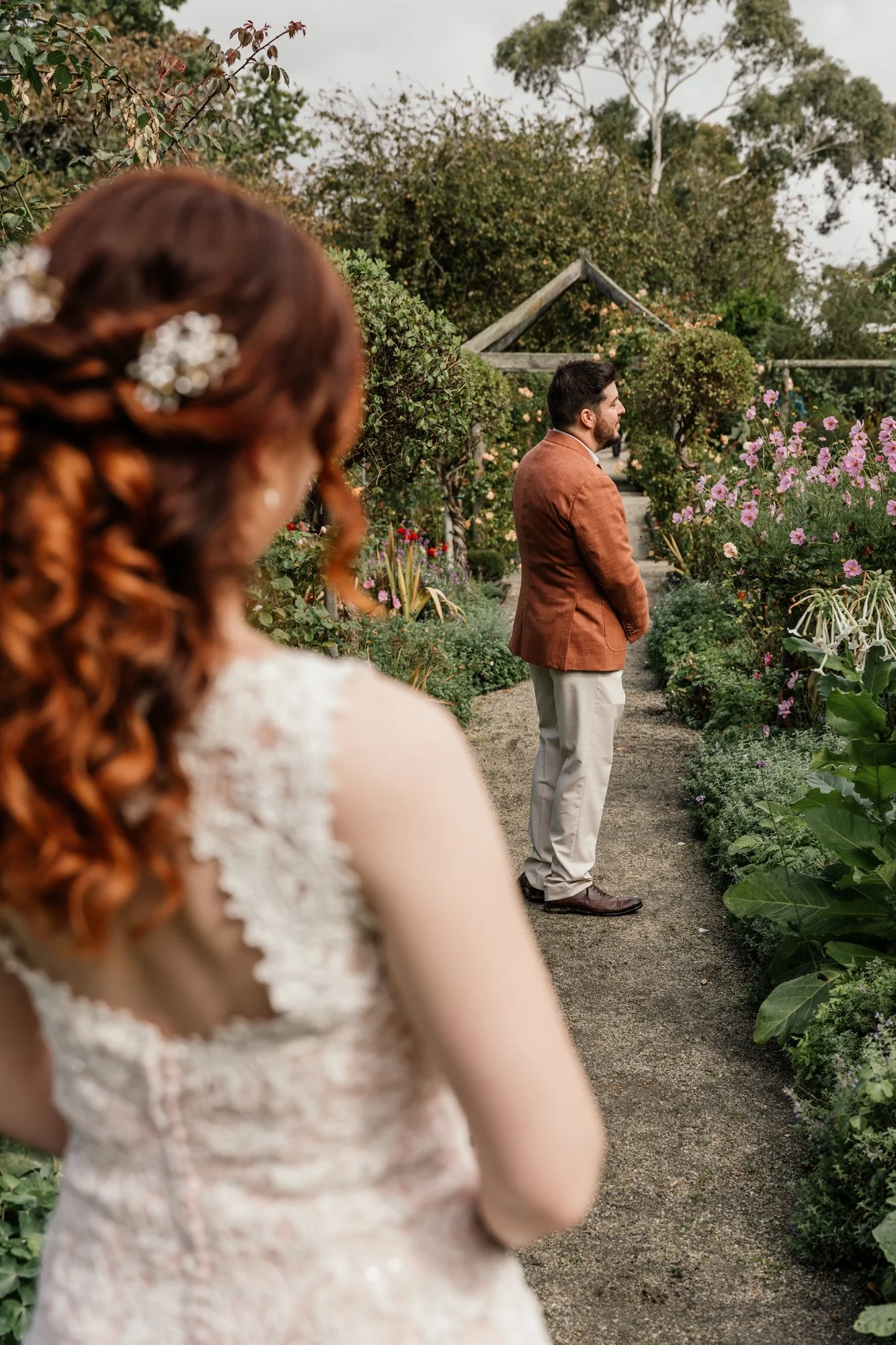A woman with curly red hair and a dress with lace details stands in the foreground. A man with dark hair and a beard, wearing a brown blazer and light-colored pants, is standing a few feet away, looking to the right, surrounded by lush greenery and p