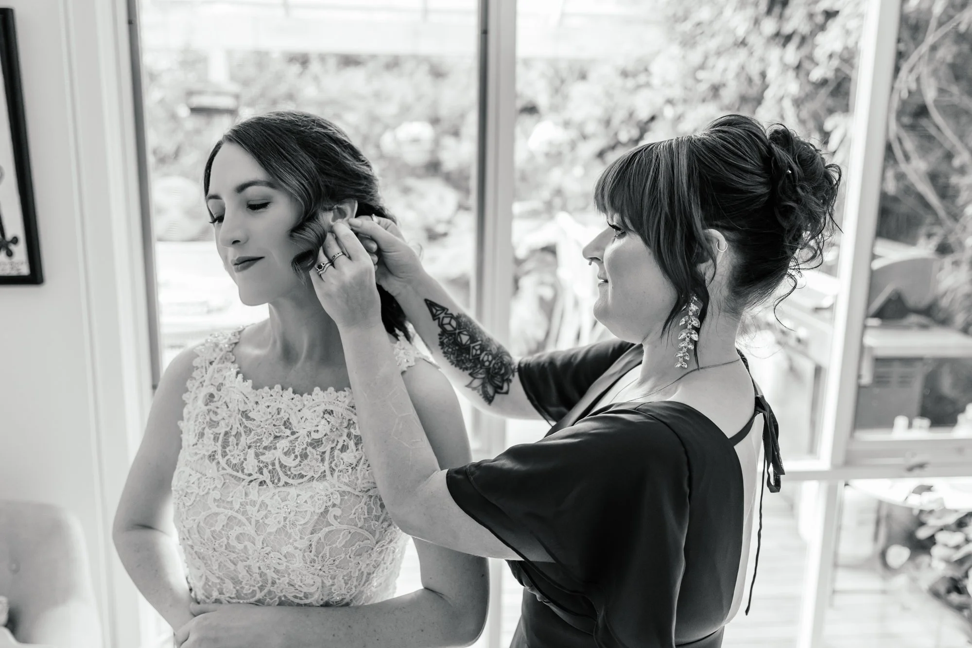 A woman in a wedding dress is having earrings put on by another woman with dark curly hair, large earrings, and tattoos on her arm, in a bright room with large windows.