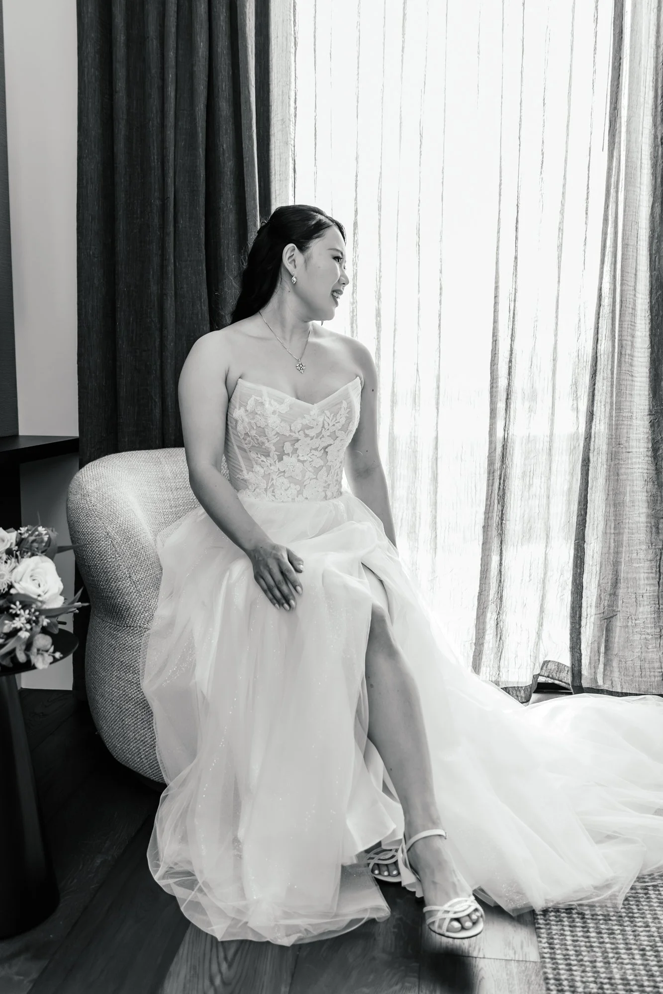 Black and white photo of a woman in a wedding dress sitting on a chair by a window with curtains, smiling and looking to the right, wearing jewelry and high-heeled shoes.