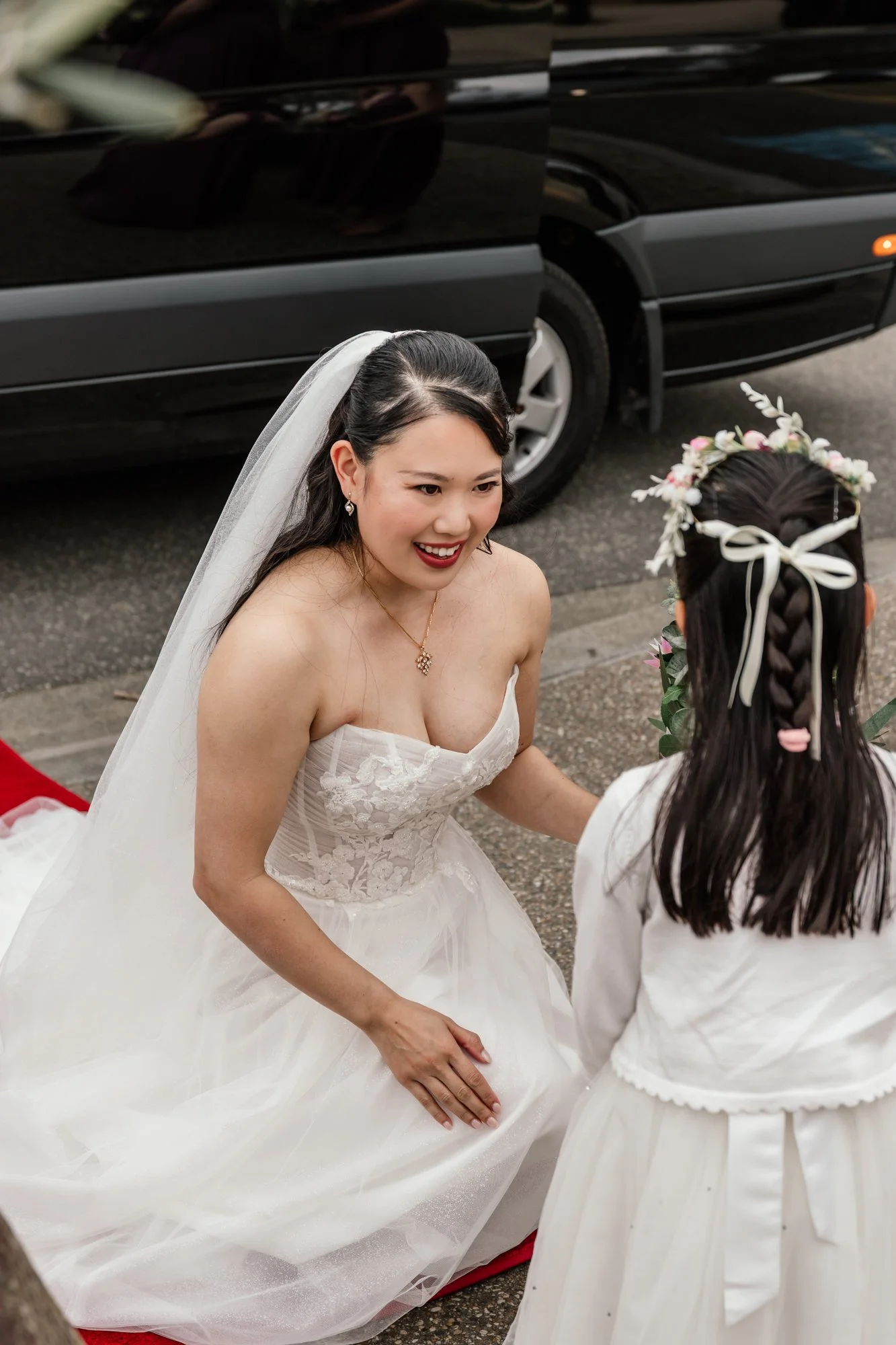 A bride in a white wedding dress with lace details on the bodice, wearing a veil, and smiling as she kneels on a red carpet, engaging with a young girl who has a floral headpiece, during an outdoor wedding ceremony.