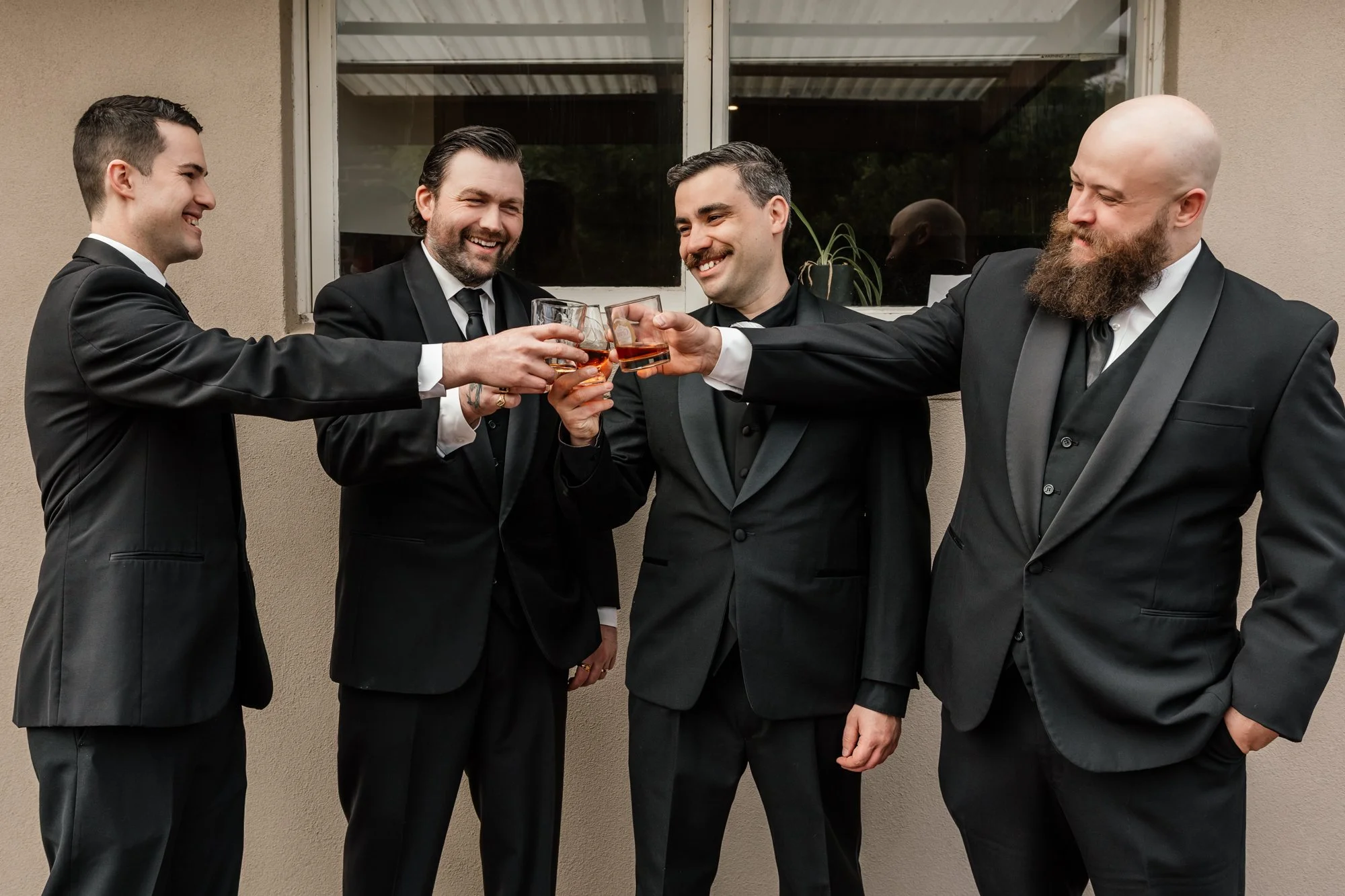 Four men in suits clinking glasses with drinks outside near a window.