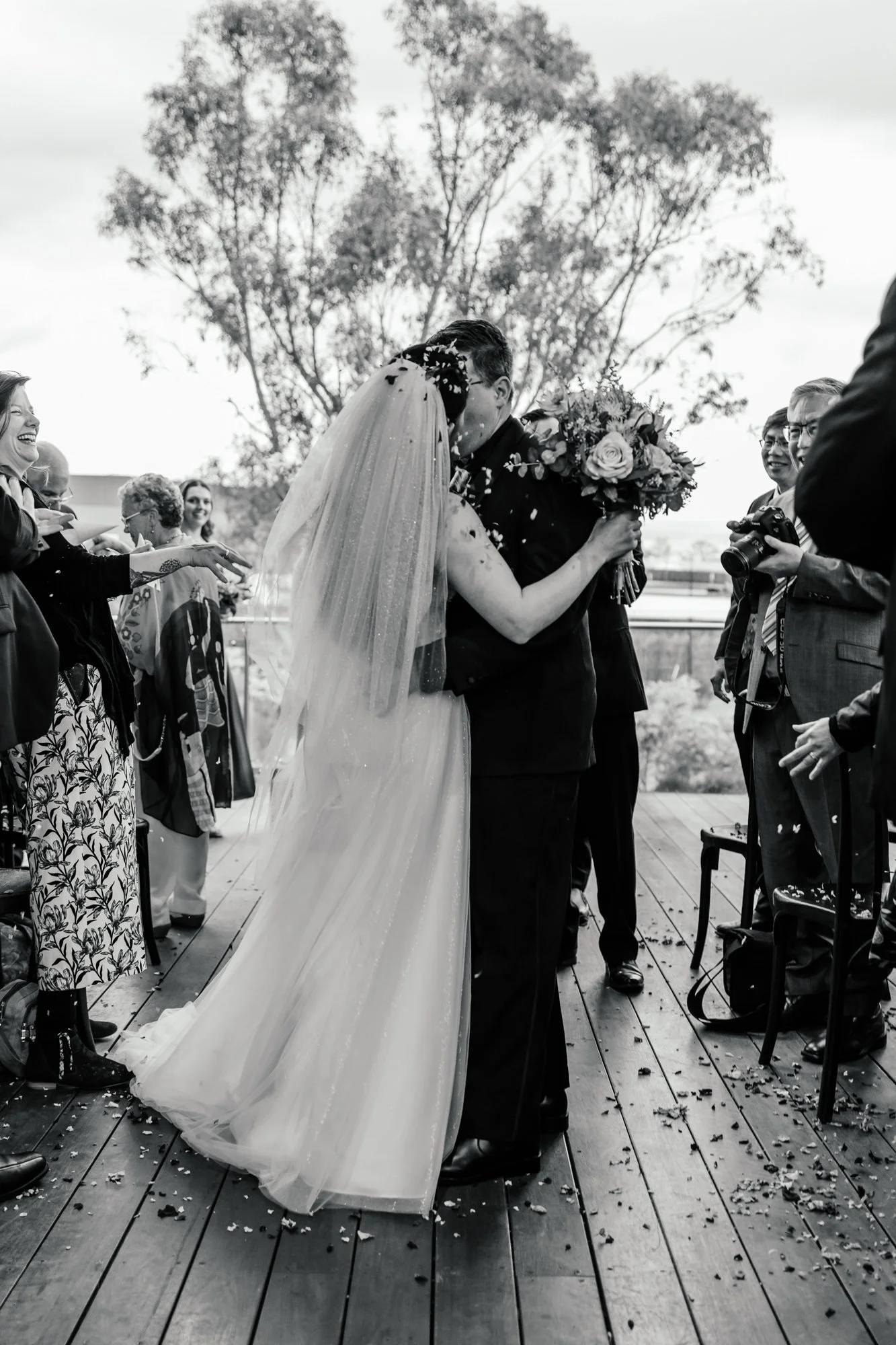 A black and white photo of a wedding celebration where the groom and bride are sharing a kiss. The bride is wearing a wedding dress and veil, holding a bouquet of flowers. Guests are gathered around, smiling and cheering, some throwing confetti on th