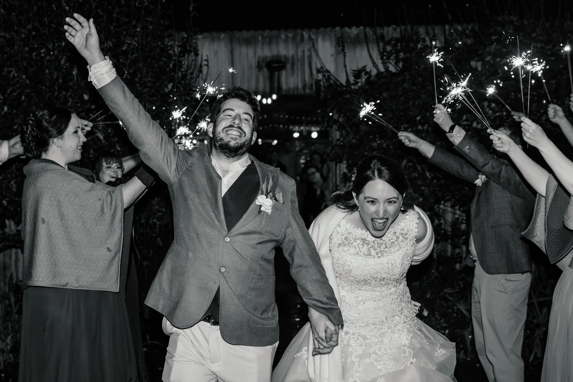 A couple in wedding attire holding hands and smiling, surrounded by people holding sparklers at night outdoors.