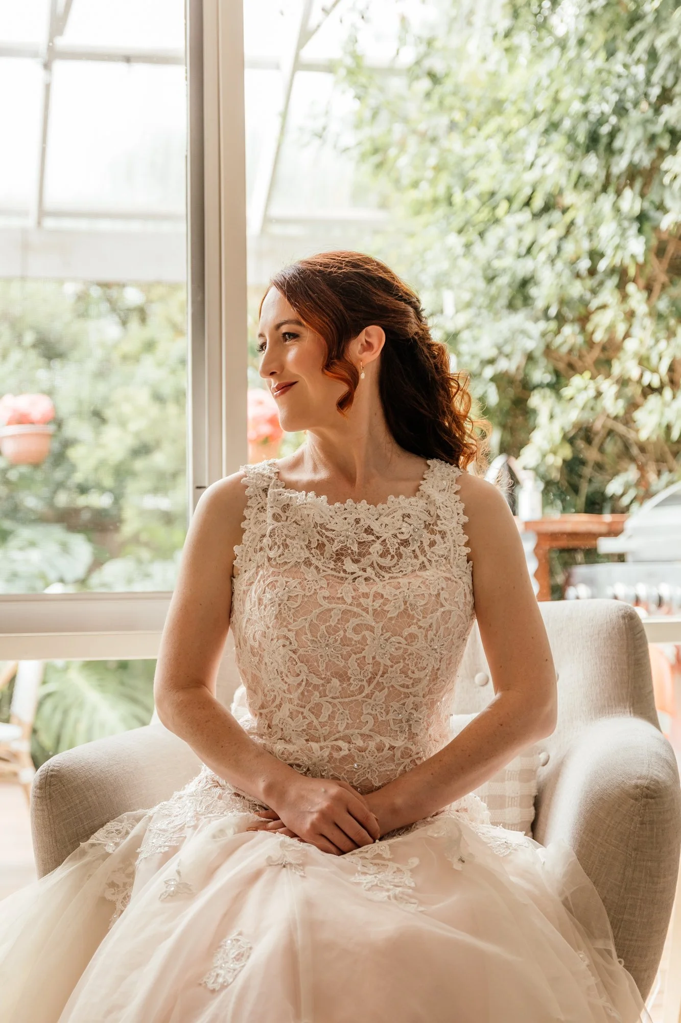 A woman in a wedding dress sitting on a beige chair by a large window with greenery outside, smiling and looking to the side.