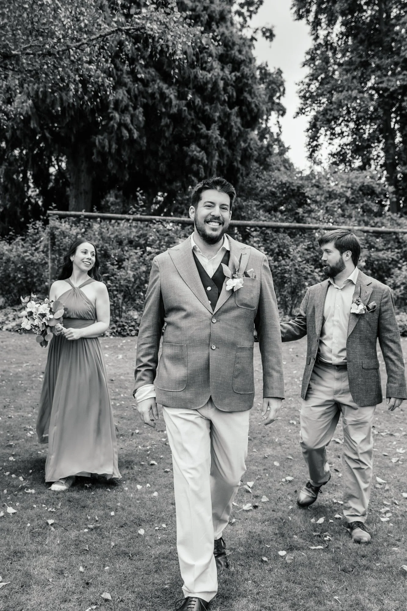 A black and white photo of three people dressed in formal attire outdoors, walking on grass with leaves scattered on the ground and trees in the background. The man in the middle is smiling, wearing a suit, and leading the group. The woman on the lef